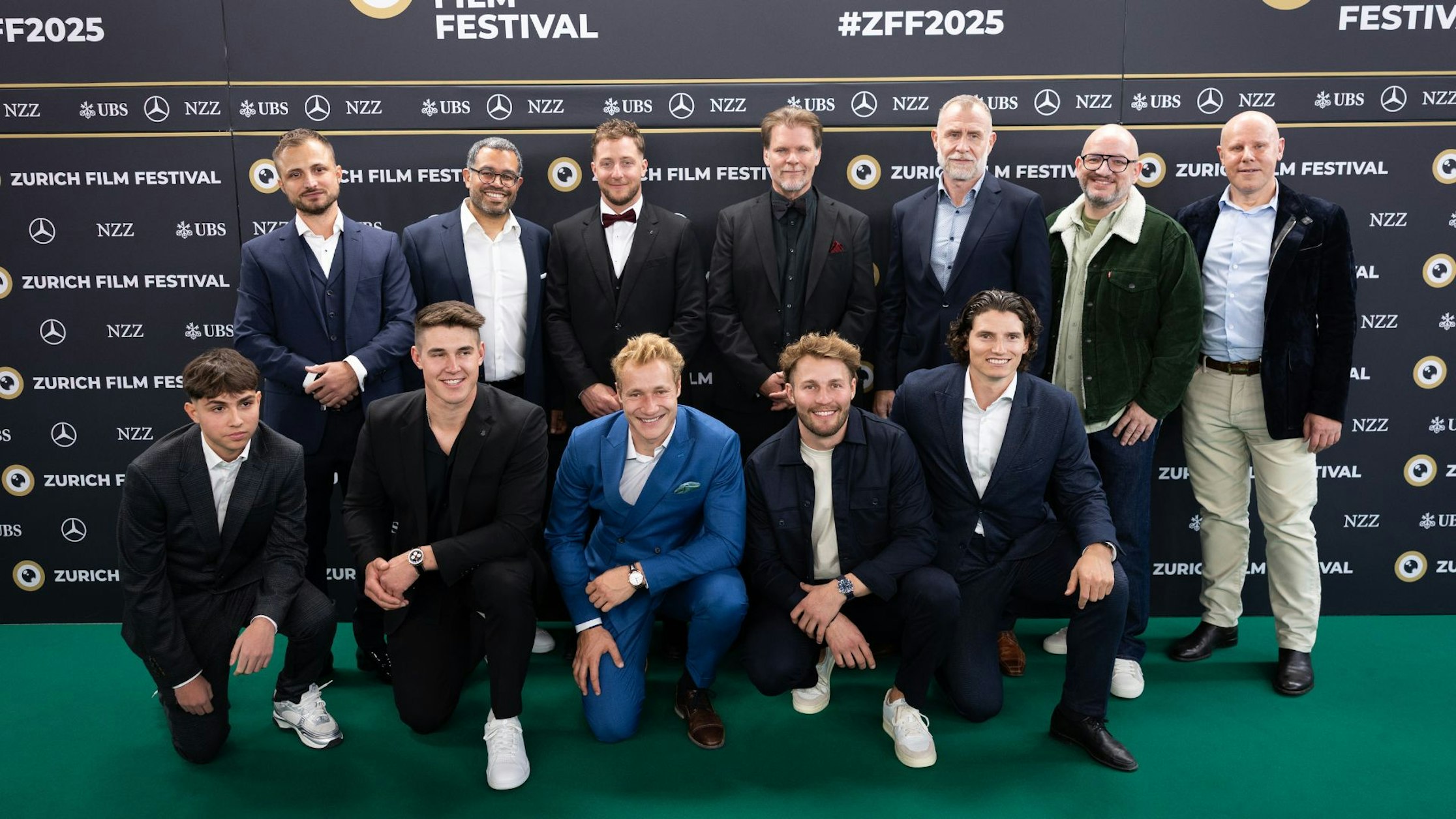 Eine Gruppe von dreizehn Männern in formellen und halbformellen Kleidern posiert gemeinsam auf dem grünen Teppich des Zurich Film Festival vor einem Hintergrund mit Logos und der Aufschrift "ZFF2025".