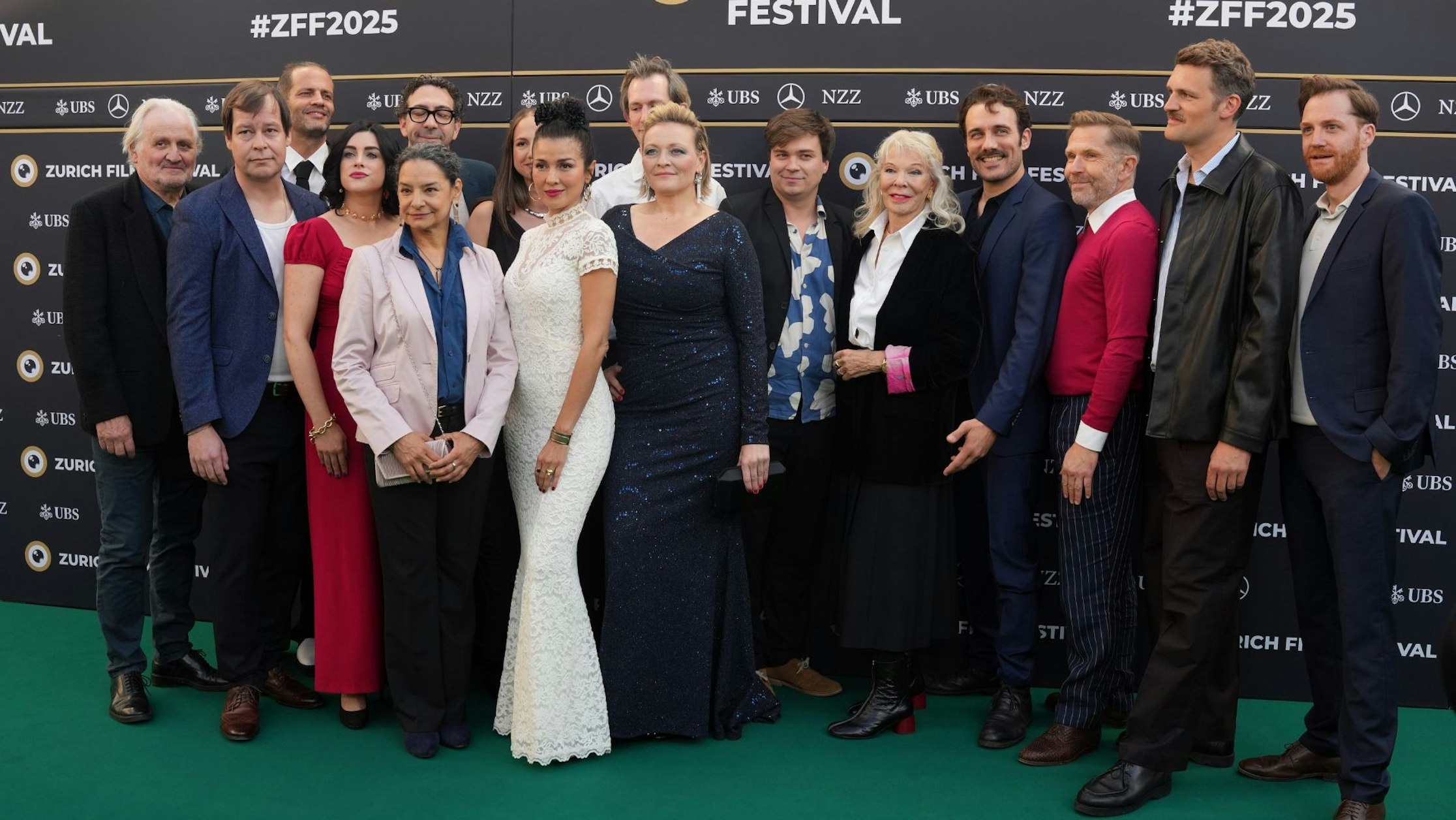 Eine Gruppe von formell gekleideten Personen steht auf dem grünen Teppich des Zurich Film Festival zusammen und posiert für ein Foto vor einer Markenkulisse.