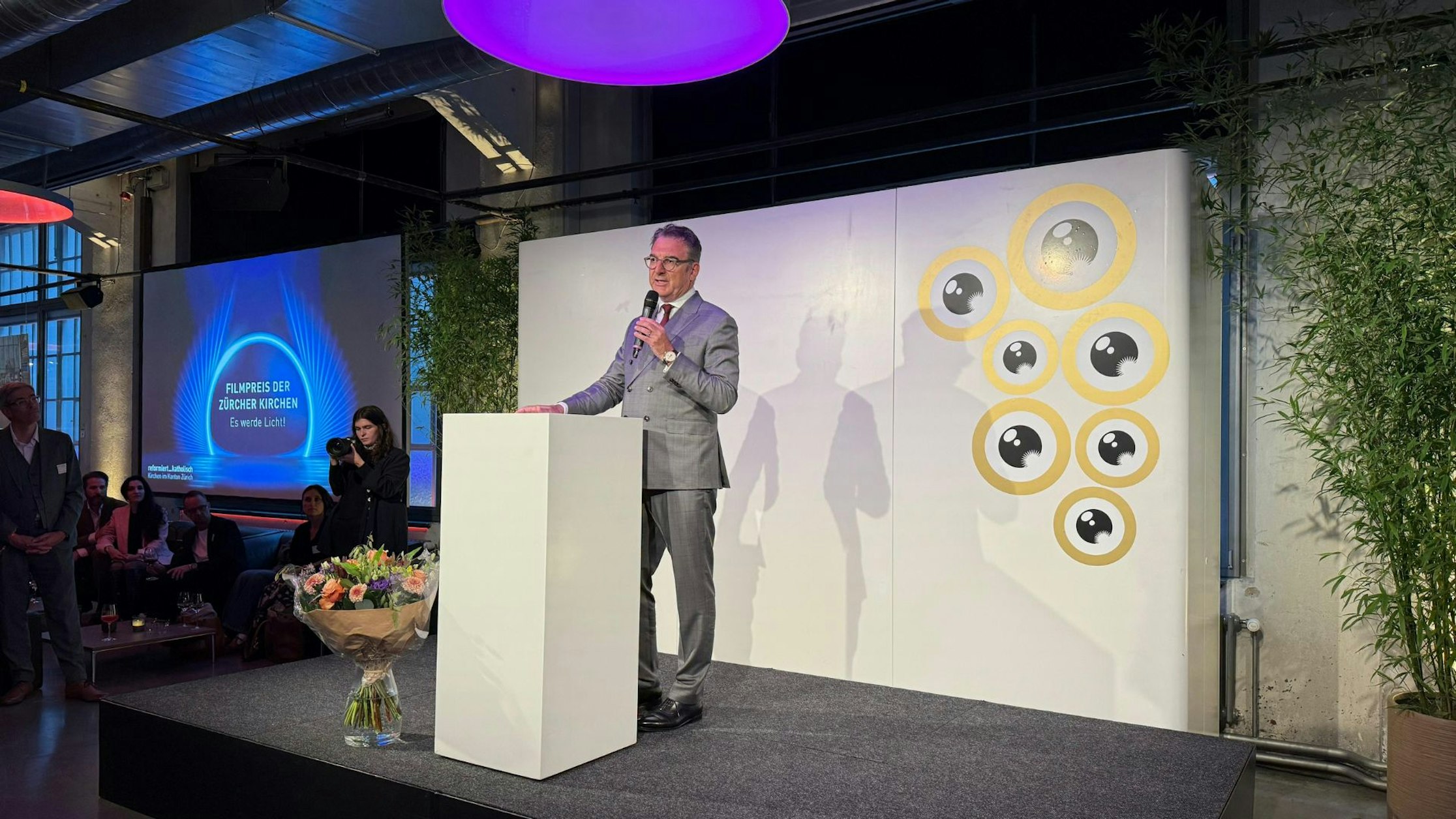 Ein Mann in einem grauen Anzug spricht an einem Podium auf einer kleinen Bühne und hält ein Mikrofon. Hinter ihm befinden sich ein weißer Hintergrund mit kreisförmigen Mustern und ein Blumenstrauß auf dem Boden. Menschen schauen von der Seite in einem modernen Innenraum zu.