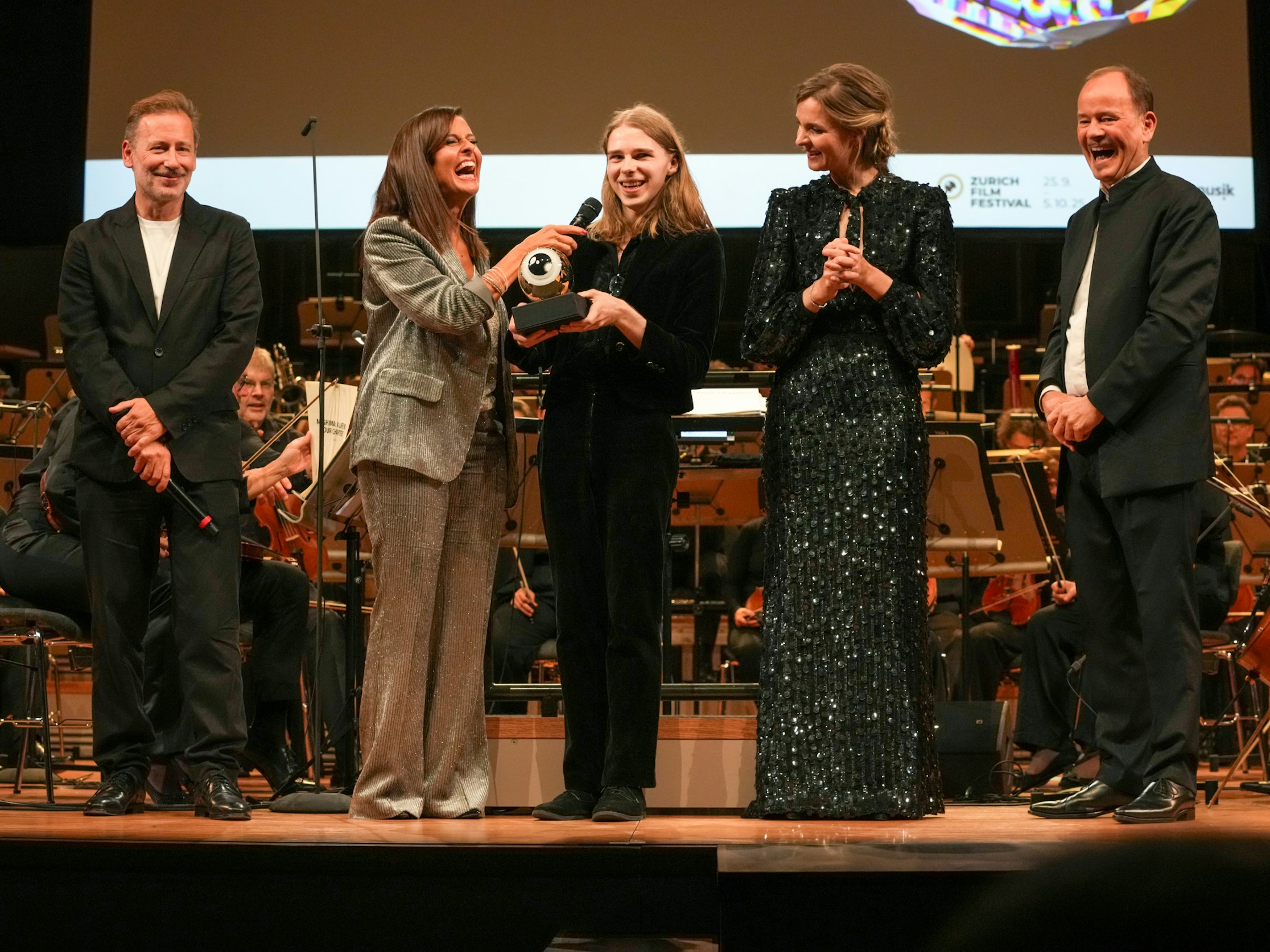 Fünf Personen stehen auf einer Bühne vor einem Orchester. Eine Frau überreicht einer lächelnden jungen Frau eine Trophäe, während die anderen lächelnd zusehen. Alle sind förmlich gekleidet und scheinen eine Leistung zu feiern.
