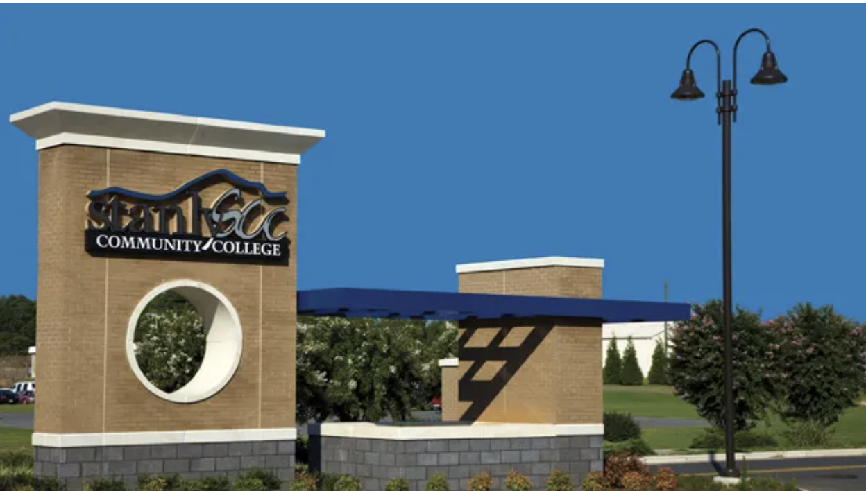 Stanly Community College entrance sign made of brick and stone, with bold lettering and a circular architectural feature. A blue awning and light post are visible near the landscaped campus entrance.
