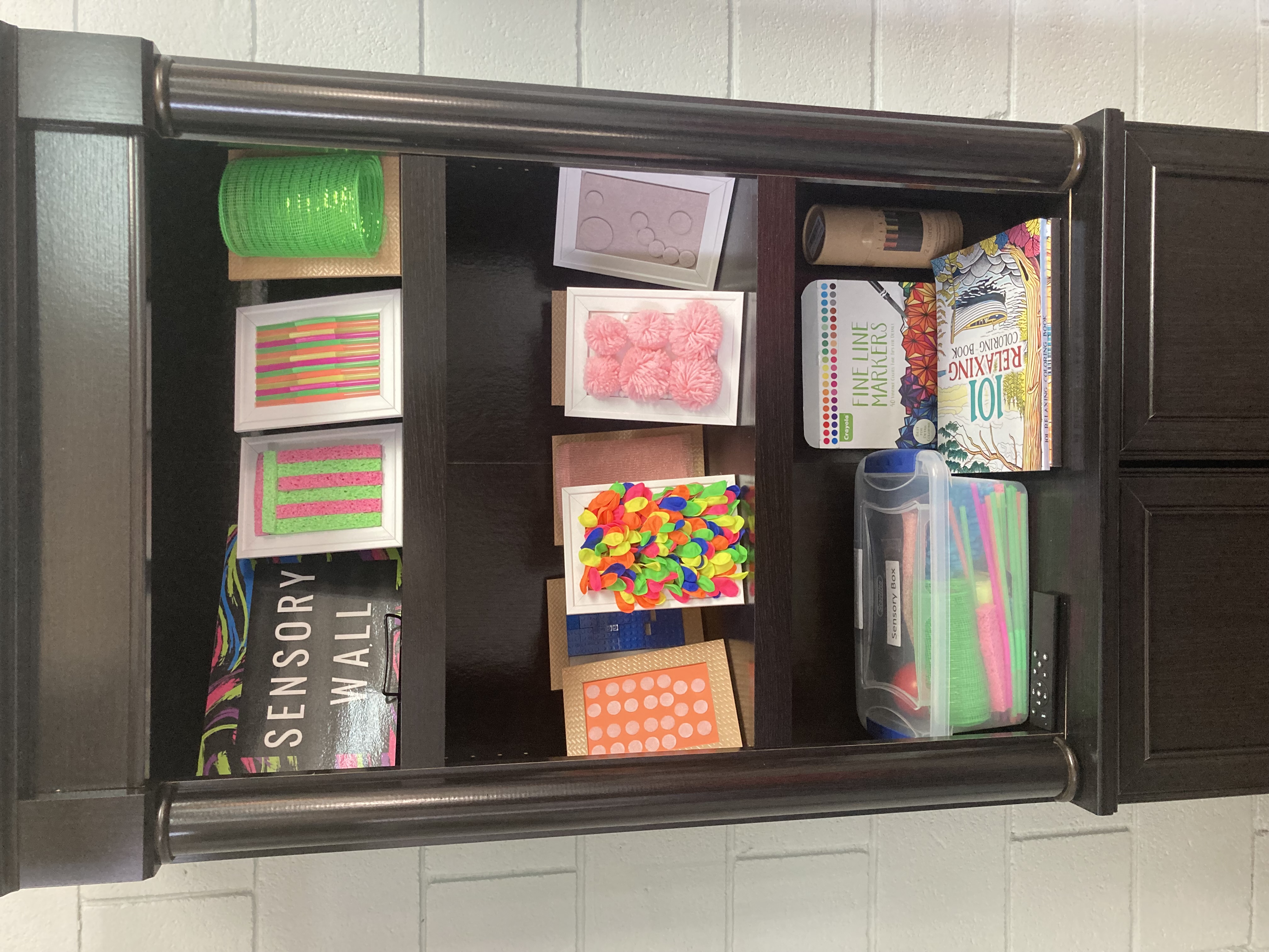 A wooden bookshelf labeled 'Sensory Wall' displaying colorful tactile sensory boards, a bin labeled 'Sensory Box' with fidget items, fine line markers, coloring books, and a sensory roller.