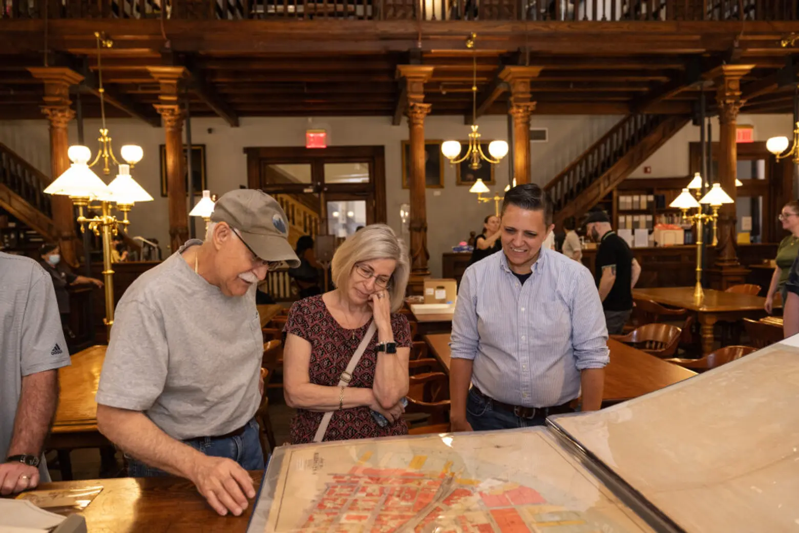 Three visitors in a wood-paneled library reading room examine a large historic map on a table.