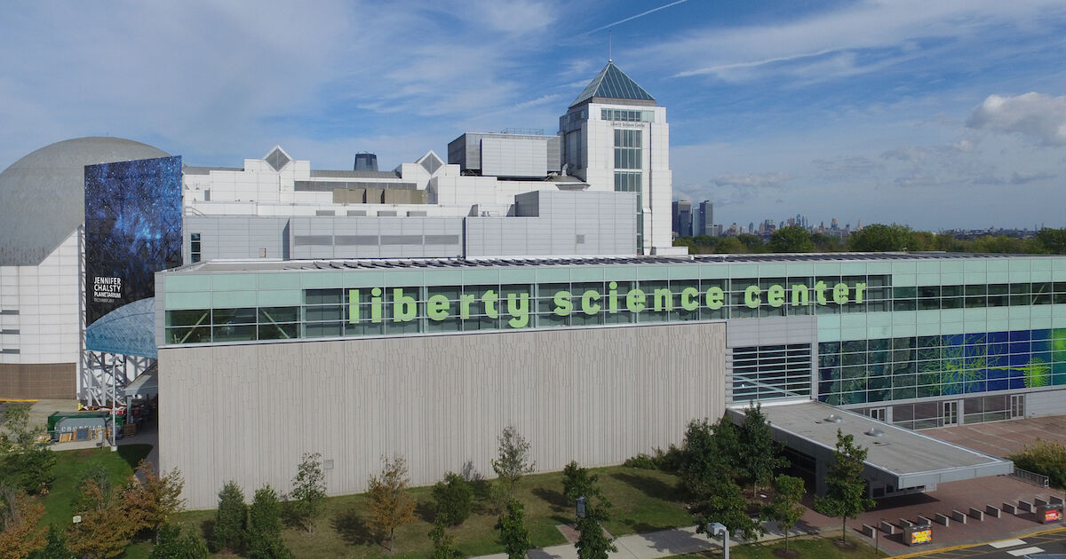 Exterior of Liberty Science Center with a long glass façade and large green letters reading “liberty science center,” viewed on a clear day with trees and walkways in front.