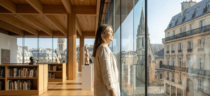 Une femme debout dans une galerie lumineuse, regardant par une grande baie vitrée sur des bâtiments historiques et une église en arrière-plan.