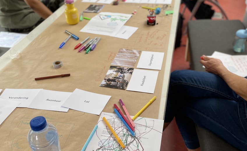 Groepje mensen en kinderen zitten aan een tafel waar papier en stiften op liggen.