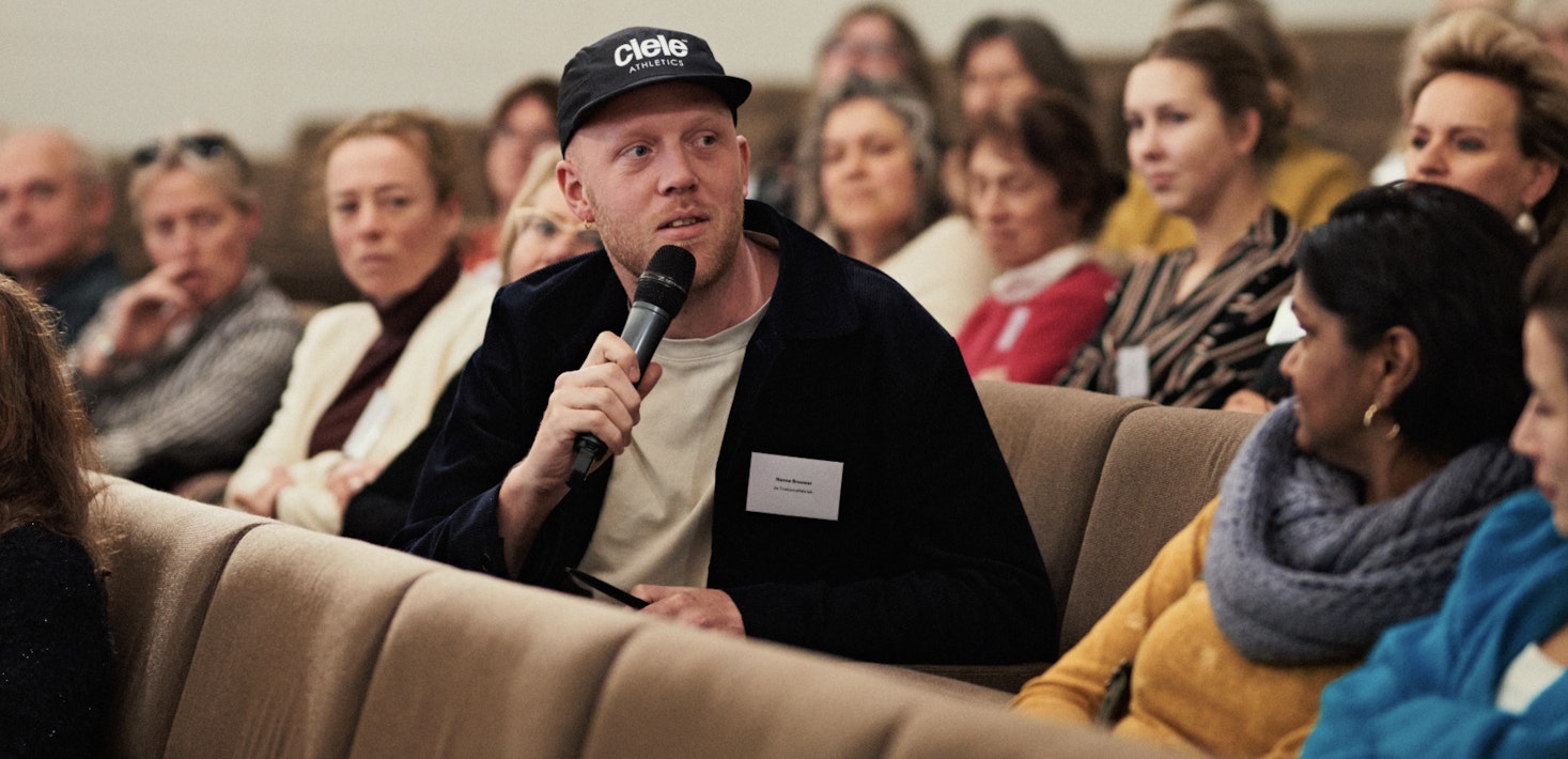 Foto van Nanne Wytze Brouwer. Hij zit in een collegezaal tussen andere mensen. Hij praat in een microfoon en kijkt naar rechts. Hij draagt zwarte kleding en een zwarte pet.