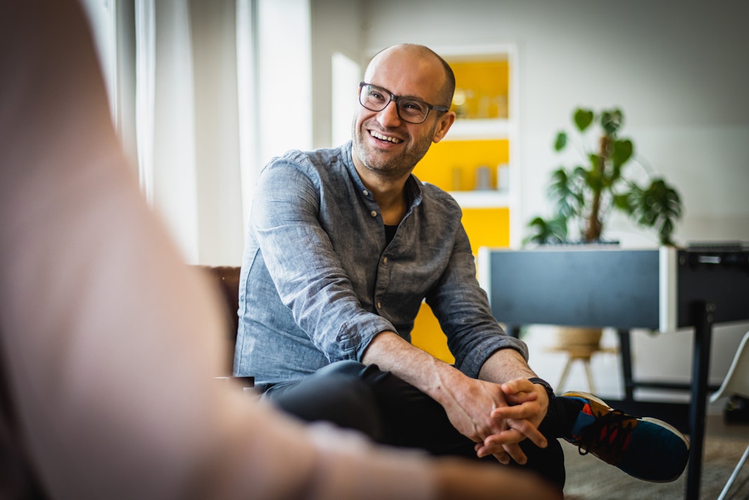 Alain zit op een stoel op kantoor en lacht naar een collega.