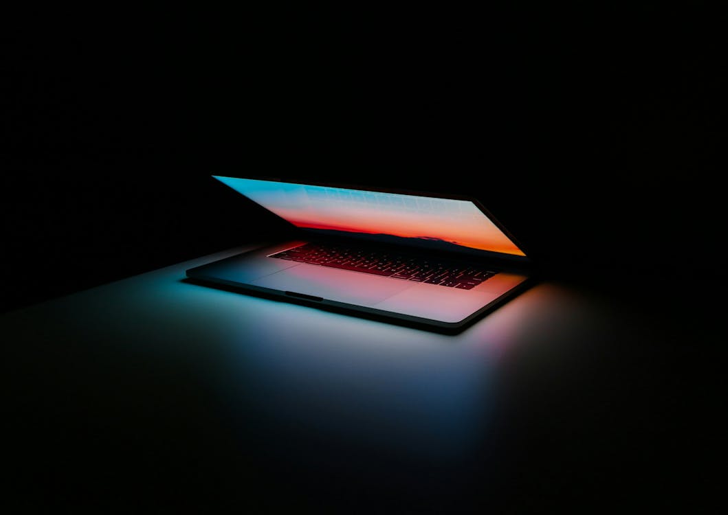 A laptop with an illuminated keyboard and screen glowing in a dark room.