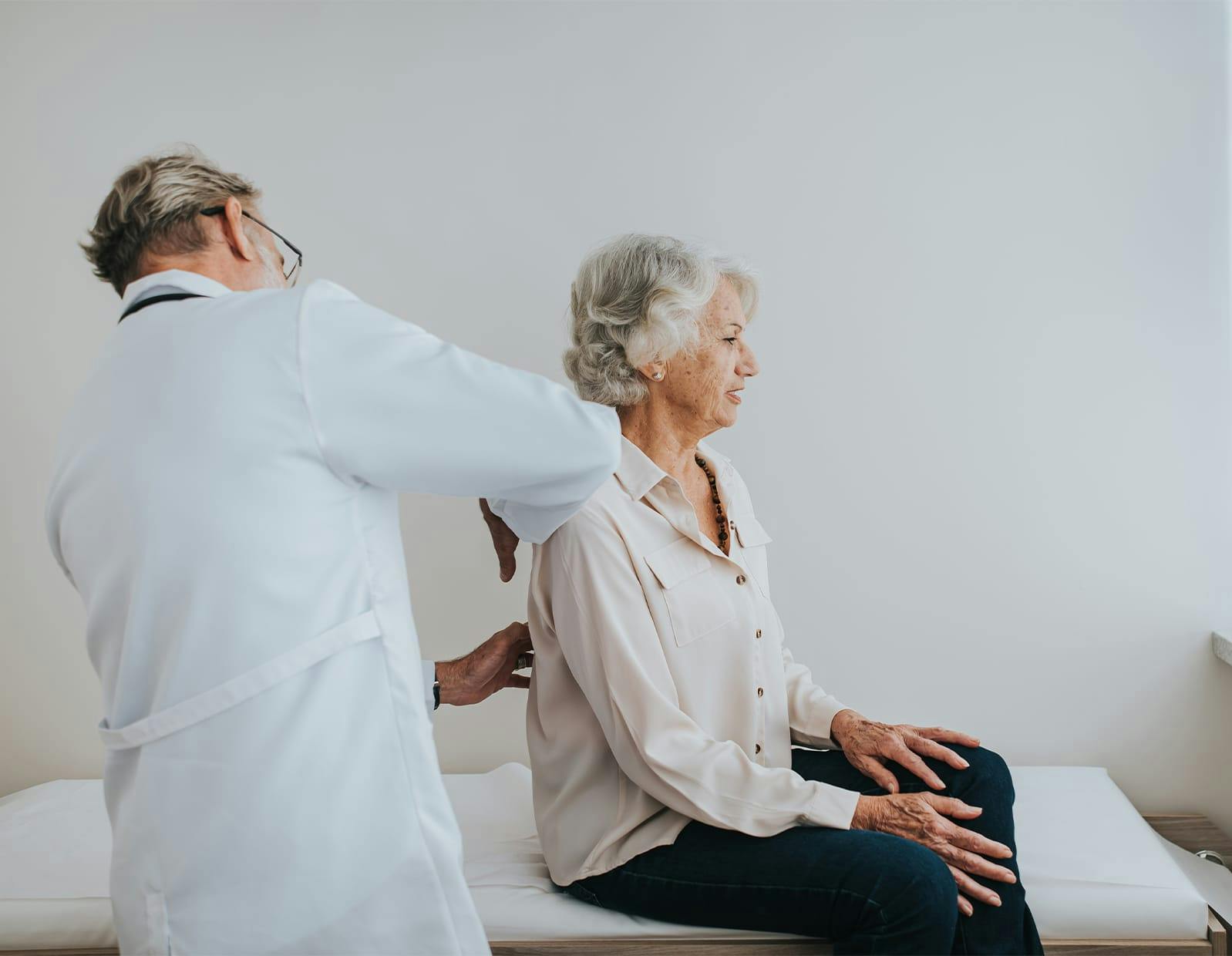 Elderly woman sitting on an exam table while a doctor performs a physical back examination