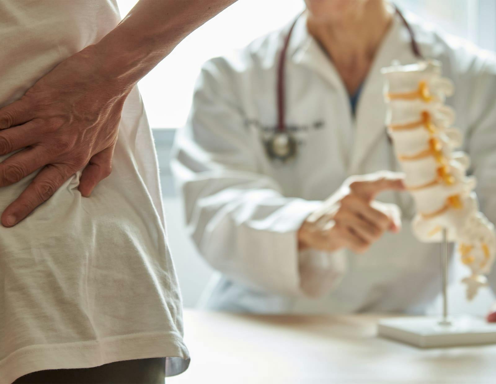 Doctor pointing to a spine model while a patient holds their lower back in pain during a consultation