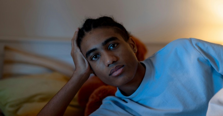 A young person lying in bed with their head on a pillow, propping up their head and appearing deep in thought, suggesting possible signs of depression.
