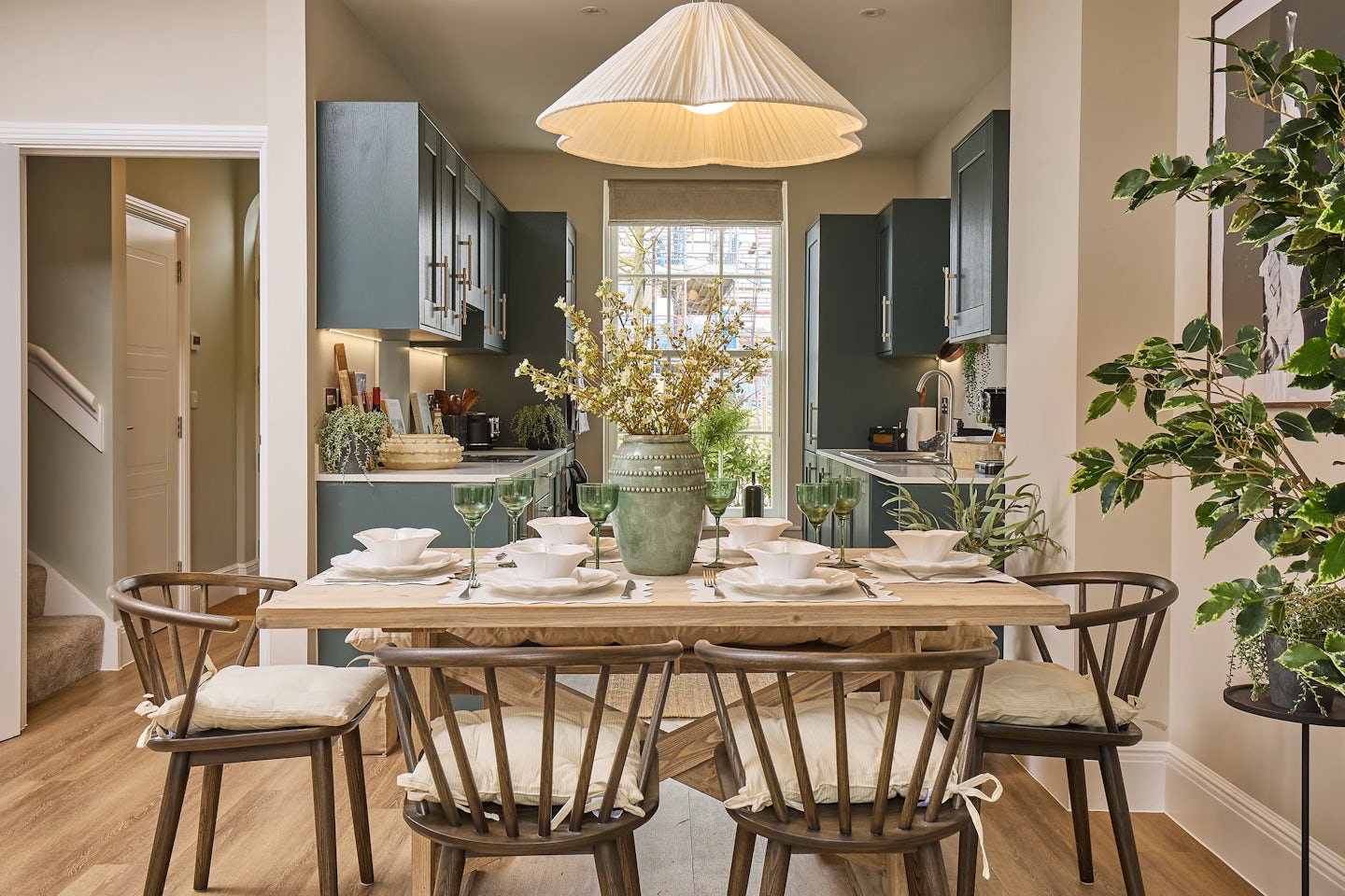 A well-decorated dining area that seamlessly transitions into a kitchen at the Burlington, Welborne. The dining table is set for six people with white plates, bowls, and green wine glasses. A green vase with yellow flowers serves as a centrepiece. The chairs have cushioned seats tied with white ribbons. In the background, the kitchen features green cabinets with gold handles, a double height sash window lets in natural light, and various kitchen appliances and decor items. A large, modern pendant light hangs above the dining table, and plants add a touch of greenery to the space.