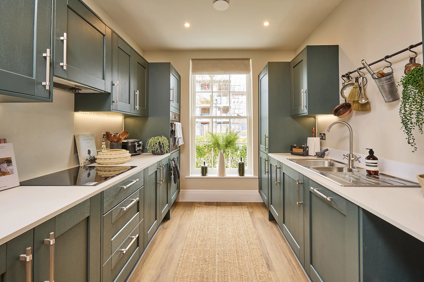 A stylish modern shaker kitchen with dark green cabinets and white countertops in a galley-style layout. The left side features an induction cooktop, woven storage baskets, a small potted plant, and a toaster. On the right side, a sink with a drying rack, a soap dispenser, and hanging kitchen utensils creates a functional space. A light-coloured rug covers the floor, and a large double height sash window at the end lets in natural light, with three potted plants placed on the windowsill.
