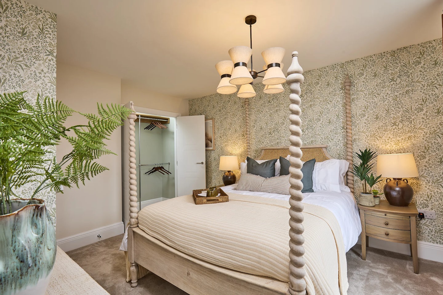 A cosy master bedroom featuring a four-poster bed with a beige blanket and multiple pillows, including dark grey and green accents. A wooden tray holding a cup, and a small plant sits atop the bed. The wallpaper behind the bed displays a floral pattern, adding elegance to the space. Two bedside tables, each with a lamp and decorative elements, flank the bed. A chandelier with five lamps hangs from the ceiling, casting a warm glow. To the right, an open closet reveals hanging clothes. A large green plant in a decorative pot sits on a dresser to the left, enhancing the inviting atmosphere with natural touches.