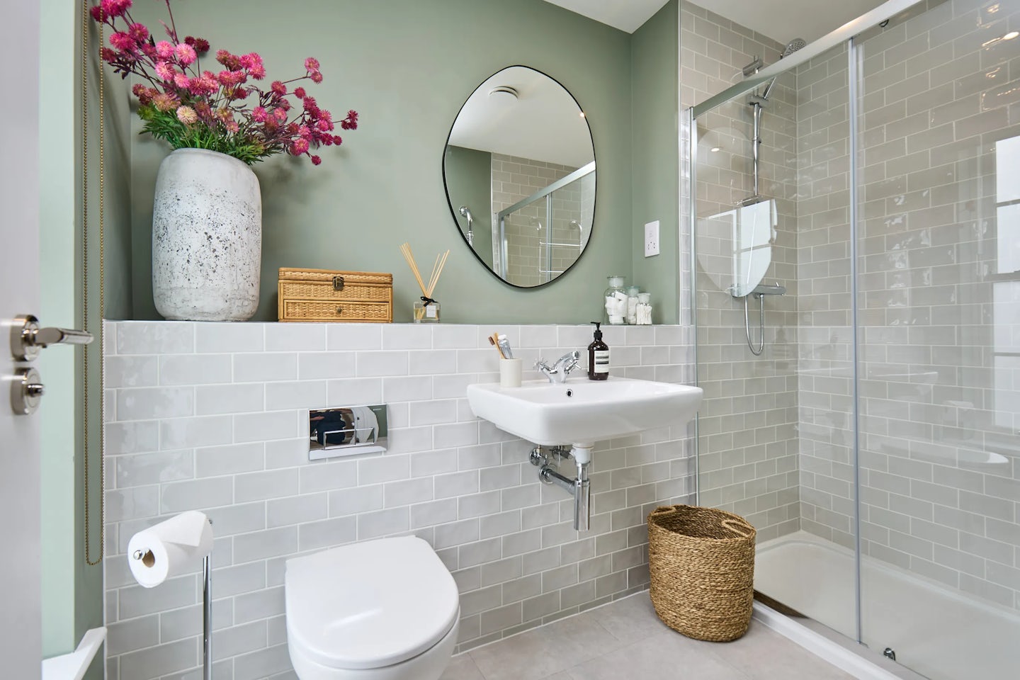 A modern bathroom featuring a white toilet with a wall-mounted flush button. To the right, a sleek white sink holds a soap bottle, with a round mirror mounted above on a green wall. A glass-enclosed shower with a rainfall showerhead and built-in shelf. Above the toilet, a decorative shelf holds a vase with pink flowers, a wicker box, and a reed diffuser. The walls have light grey subway tiles, and the floor is covered in light-coloured tiles.