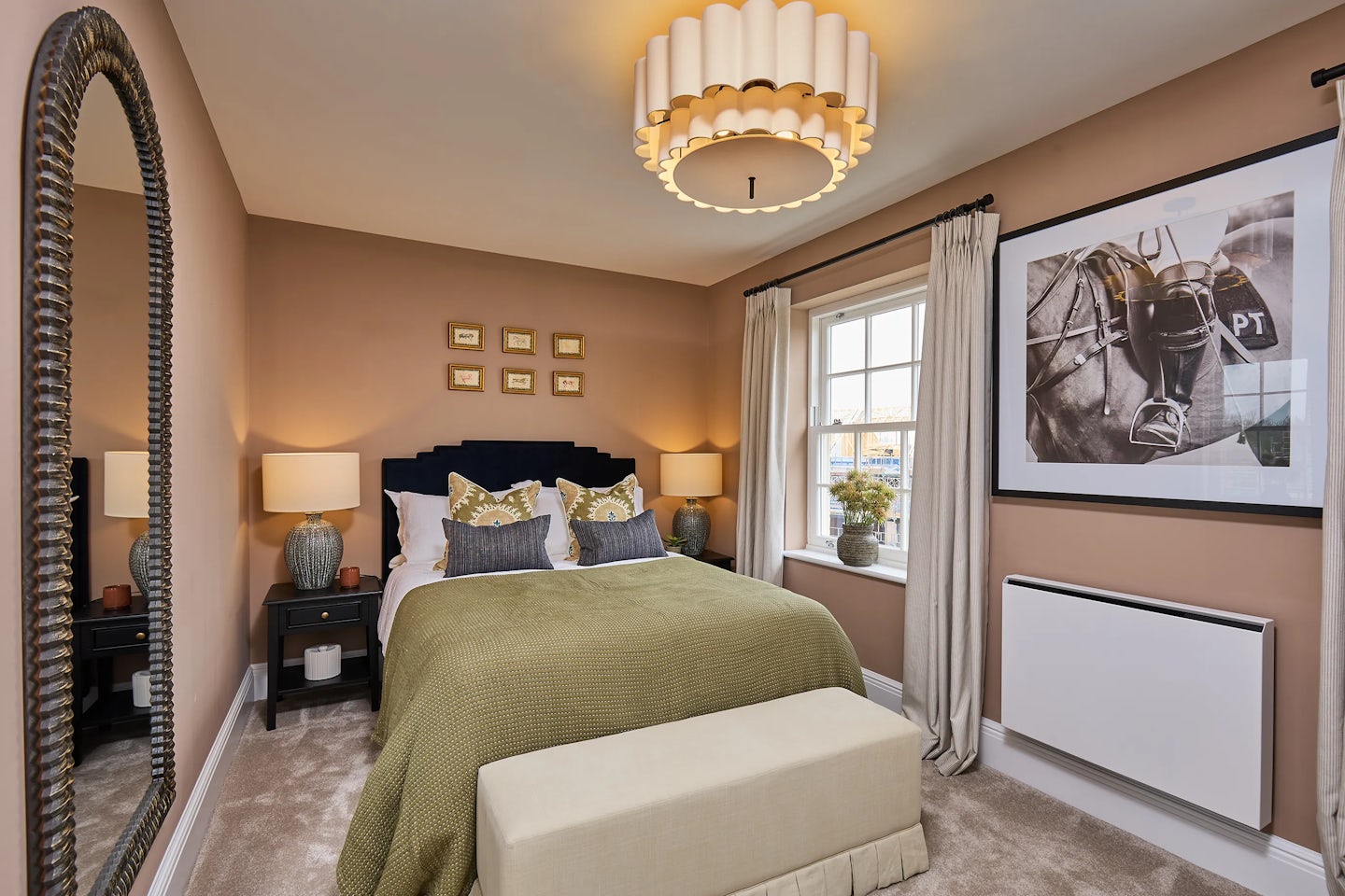 A cozy bedroom with a neatly made double bed featuring white linens and a soft throw blanket. A bedside table with a lamp sits to the right of the bed. Large windows with floor-length curtains allow natural light to fill the space. A framed picture hangs above the bed, adding a decorative touch.