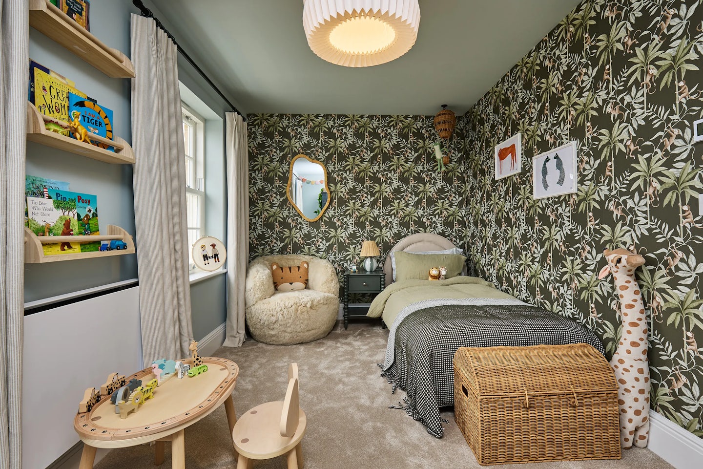 Playful children's bedroom with jungle-themed wallpaper featuring monkeys and palm trees. The room includes a single bed with green bedding, a cosy faux-fur chair, wicker toy chest, and wooden table with animal toys. Bookshelves line the wall beside a window with striped curtains, and a soft giraffe toy stands in the corner.