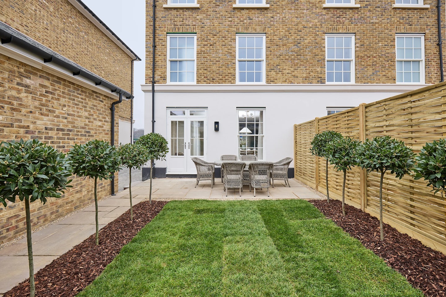 Neatly landscaped rear garden with a central lawn, flanked by symmetrical rows of small, round bay trees bordered with bark mulch. An outdoor dining set sits on a stone patio against the backdrop of the Burlington at Welborne, a traditional-style brick townhouse with large sash windows and French doors.