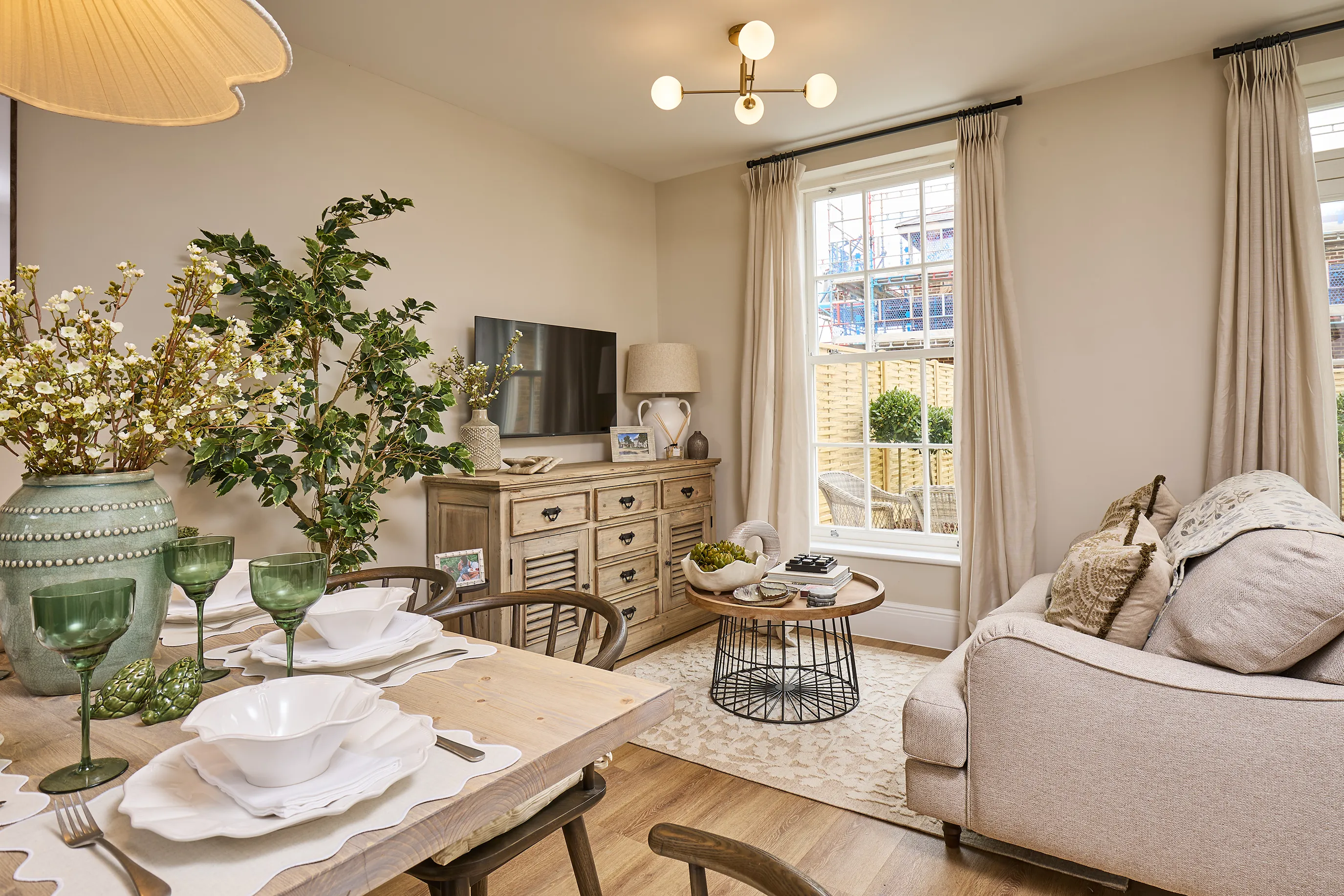 Cosy, well-lit living and dining area in the Burlington at Welborne - featuring neutral tones, a pale sofa, wooden dining table set for four, and rustic-style sideboard. Decorative elements include green glassware, vases of artificial flowers, and framed photos. Natural light streams in through tall sash windows with cream curtains, offering a view of the garden and patio beyond.