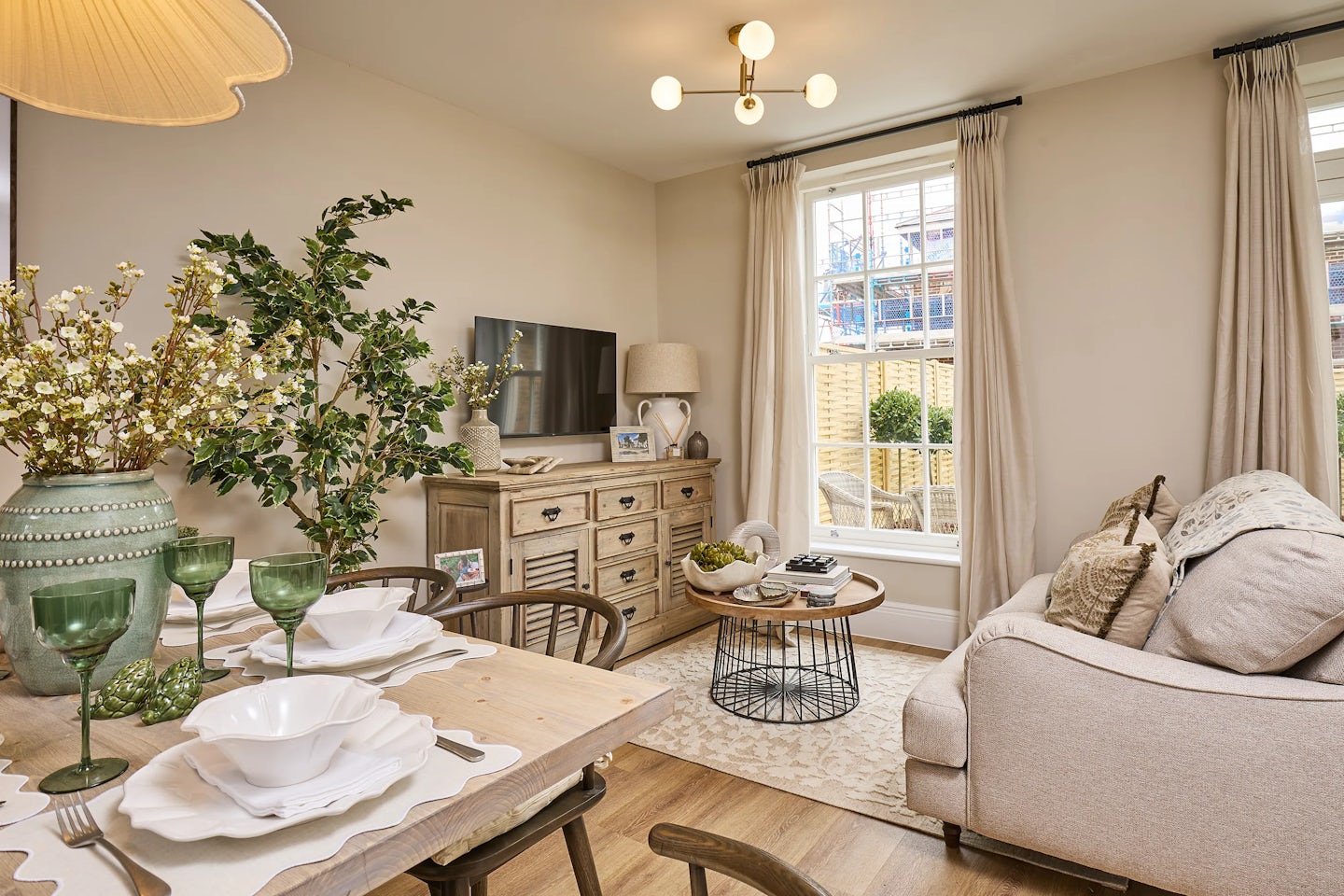 Cosy, well-lit living and dining area in the Burlington at Welborne - featuring neutral tones, a pale sofa, wooden dining table set for four, and rustic-style sideboard. Decorative elements include green glassware, vases of artificial flowers, and framed photos. Natural light streams in through tall sash windows with cream curtains, offering a view of the garden and patio beyond.
