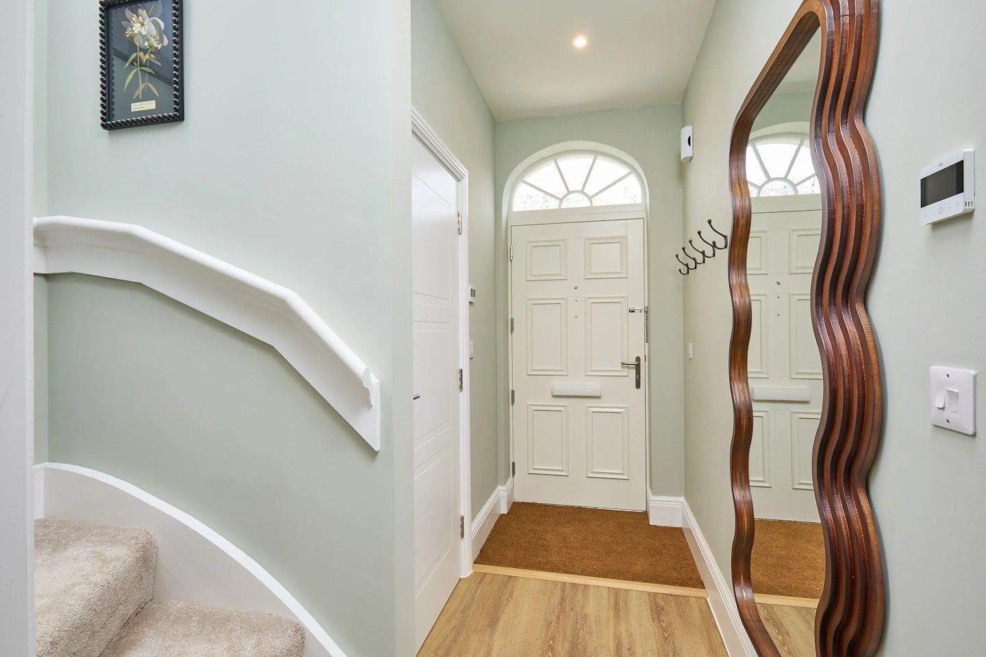 A well-lit hallway with light green walls and a wooden floor. A carpeted staircase with white railings leads to the upper level. A white front door with a circular window stands at the end of the hall. A large, wavy-framed mirror hangs on the right wall.