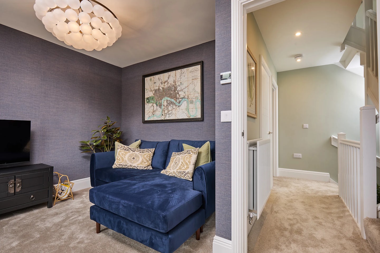 A cozy living room on the first floor of the Burlington at Welborne featuring a blue sectional sofa with patterned throw pillows. A framed map hangs on the wall above the couch, adding character to the space. A potted plant sits nearby, bringing a touch of greenery. In the background, a hallway leads to a staircase, with a radiator and a framed picture visible along the wall.