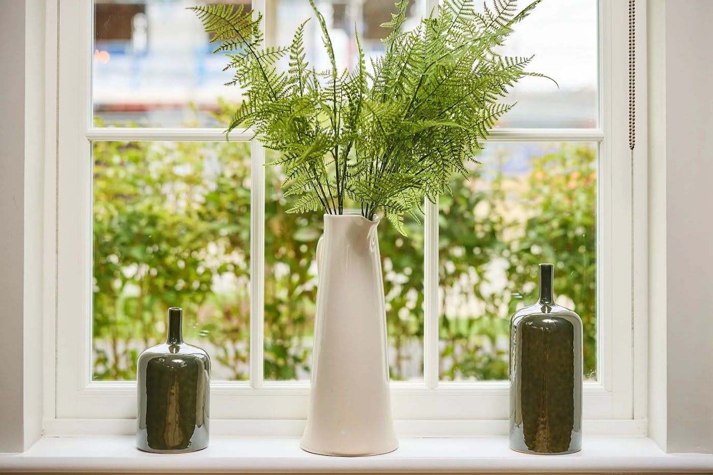 A vase with ferns sits in the middle of a white sash window with two jars on either site.