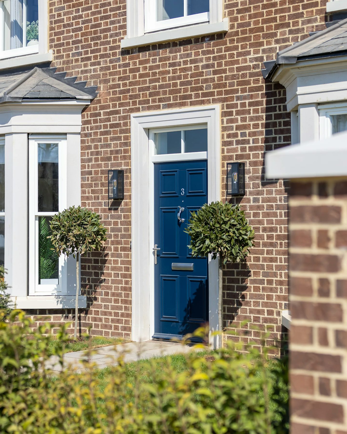 A symmetrical entrance with a blue door framed by white trim, set within a brown brick house. Two matching topiary trees in planters stand on either side. White-framed windows with shutters flank the entrance, and black lanterns are mounted on the walls.