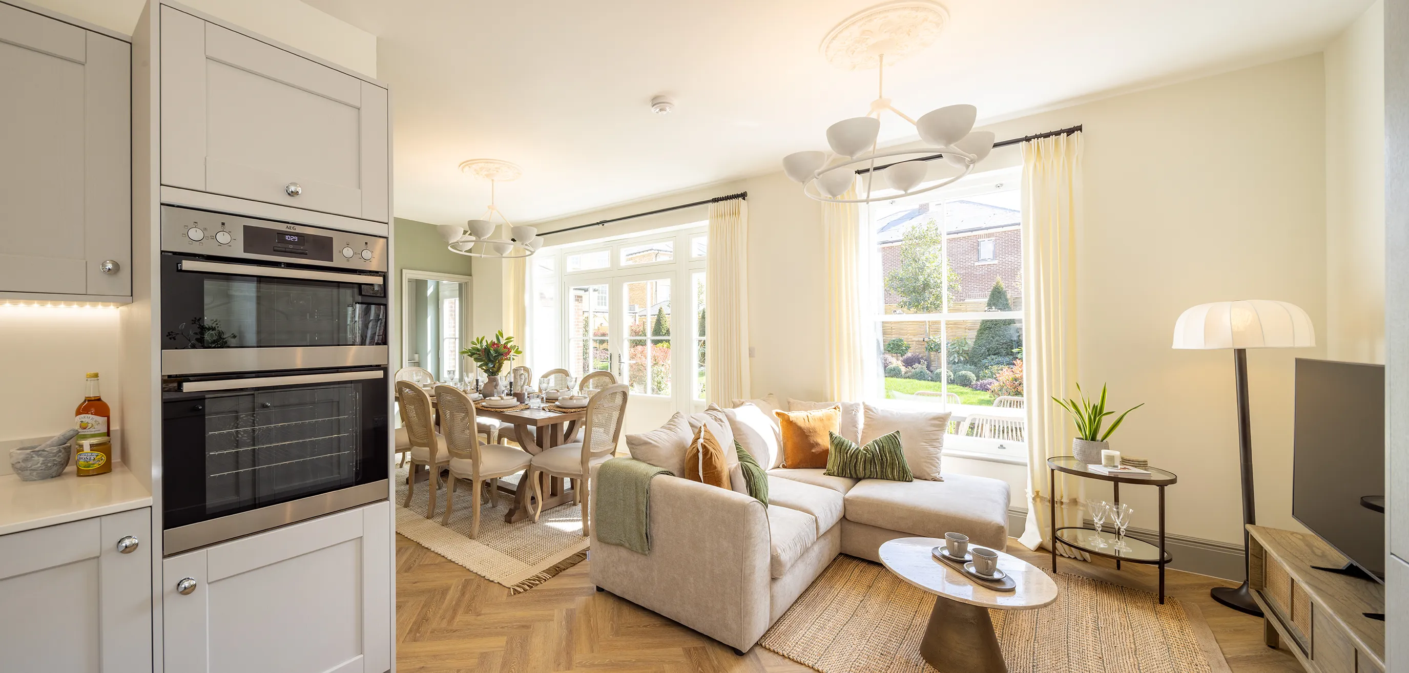 A modern open-plan living and dining area featuring a sleek kitchen with built-in appliances, a dining table set for a meal, and a cozy lounge space with a sectional sofa, coffee table, and television. Large windows fill the room with natural light, offering a view of a garden outside. The space is decorated with contemporary furniture and stylish light fixtures.