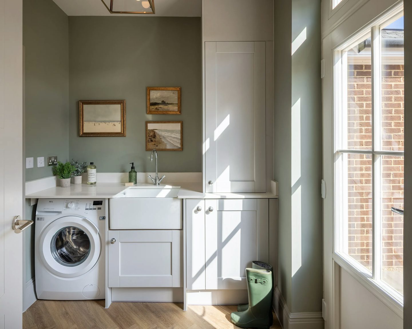A bright and modern laundry room featuring white cabinets, a wooden floor, and a large window allowing natural light to stream in. A washing machine sits beneath the countertop, next to a sink with a sleek faucet. Above the sink, three framed pictures add a decorative touch.