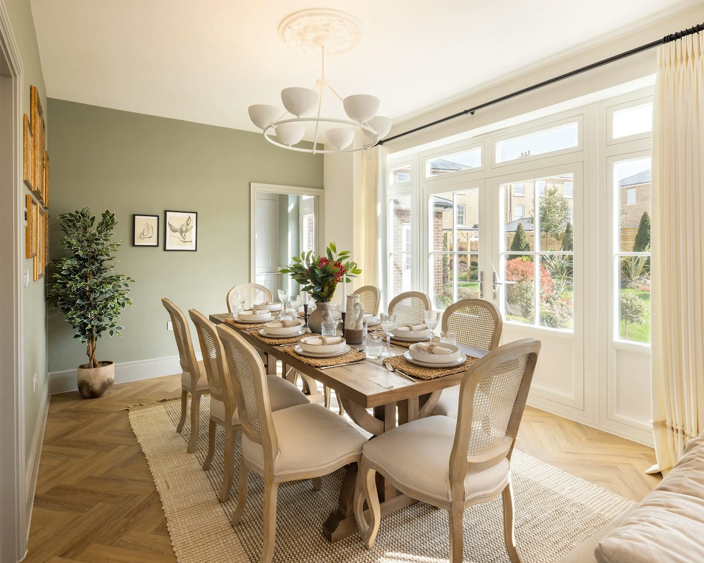 A well-lit dining area featuring a large wooden table set for a meal. The table is surrounded by eight cushioned wicker chairs and adorned with plates, glasses, cutlery, and a floral centerpiece. A large window lets in natural light and offers a view of a lush garden. The soft green walls are decorated with framed artwork, while a potted plant sits nearby. A modern chandelier hangs above.