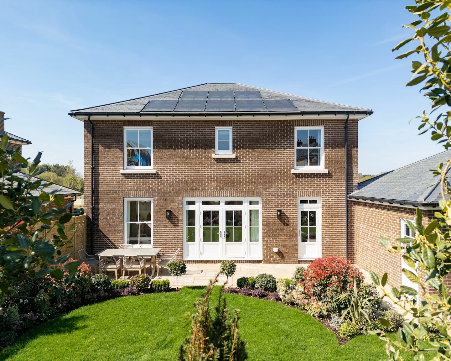 A two-story brick house with solar panels on the roof, surrounded by a well-kept garden. The symmetrical facade features large windows, including a circular window at the center. The front yard has a lush green lawn, neatly trimmed shrubs, and a patio with outdoor furniture.