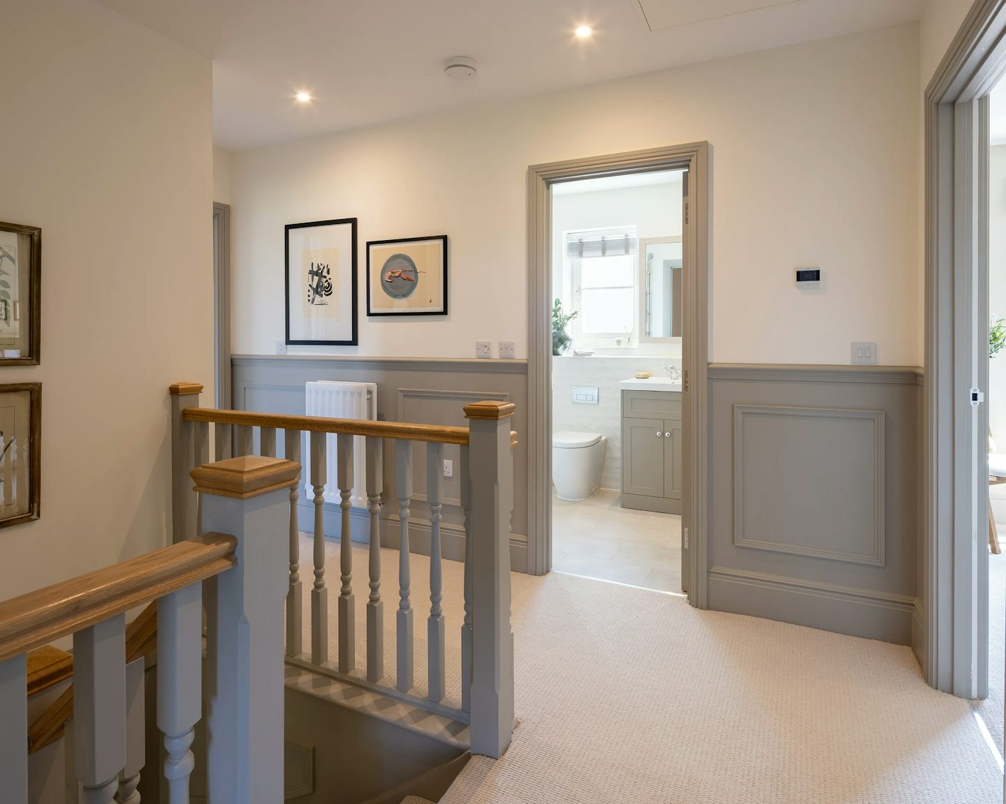 A well-lit hallway on an upper floor with a wooden staircase leading downward. The walls are painted light with contrasting grey wainscoting. Two framed pictures hang on the left wall. An open door at the end of the hallway reveals a bathroom with a toilet, sink, and a window allowing natural light. The floor is carpeted in a neutral tone, and recessed ceiling lights provide illumination.