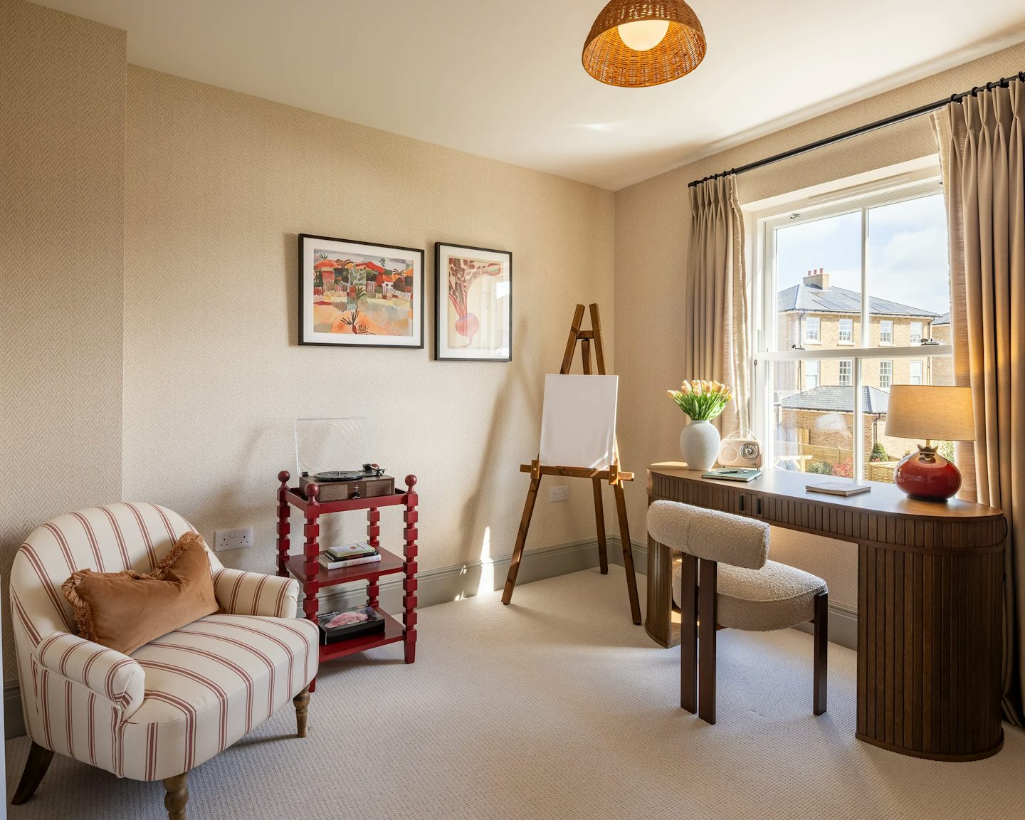 A bedroom being used as a home office and art studio. A wooden desk holds a lamp, books, and a vase with flowers. To the left, an easel with a blank canvas suggests artistic pursuits. Above it, two framed artworks decorate the wall. In the foreground, a striped armchair with a brown cushion sits beside a small red wooden shelf with a glass container.