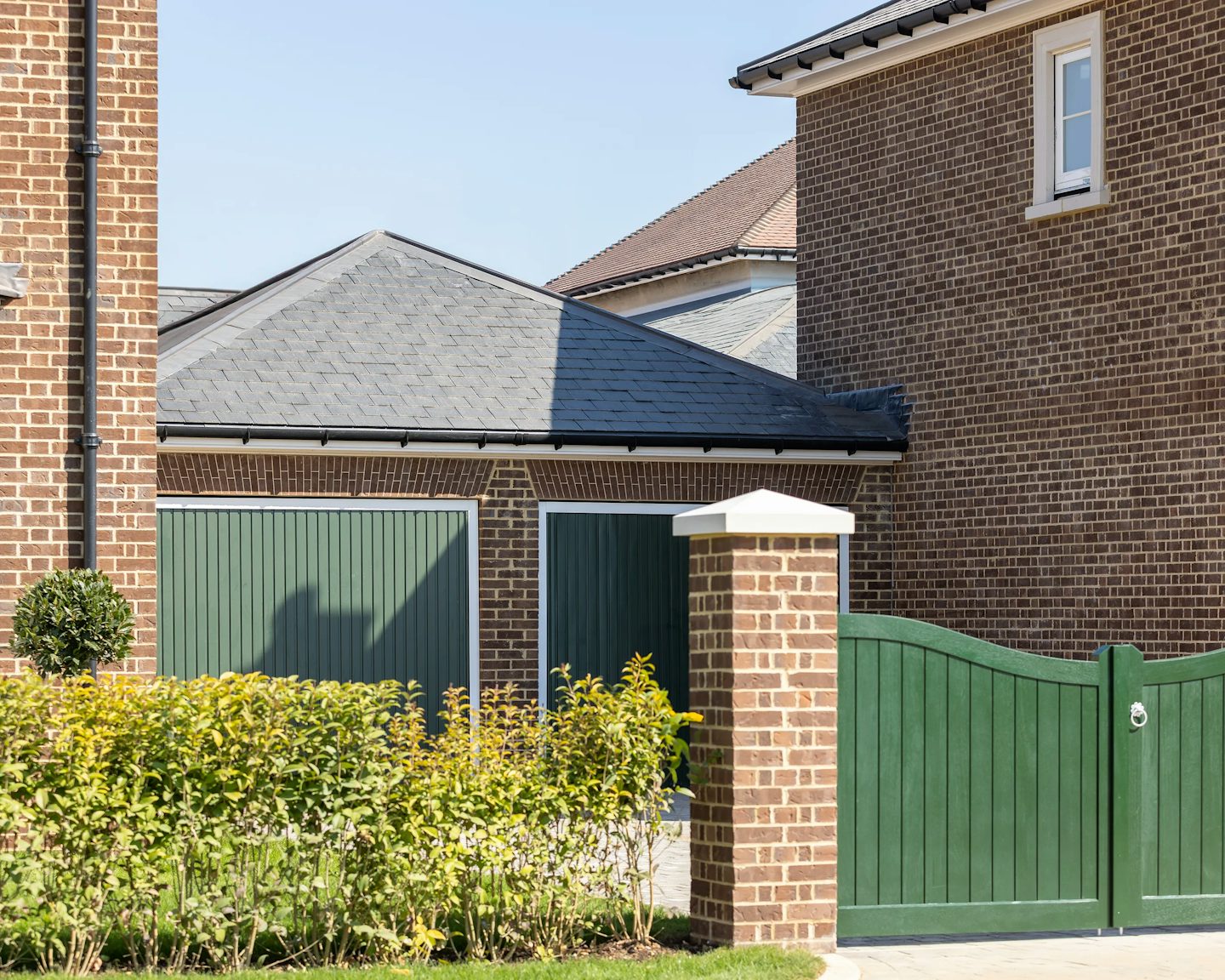 A  double brick garage with dark green doors. A green gate, supported by a brick pillar, is positioned next to the garage. A neatly trimmed hedge lines the front of the property, adding to its well-kept appearance.
