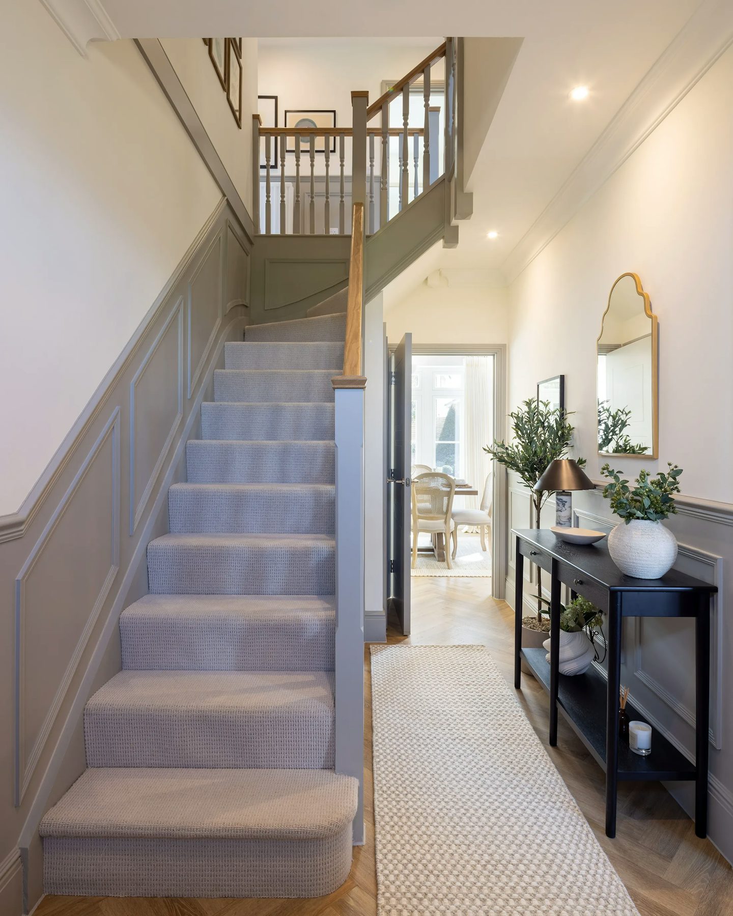 A well-lit hallway with a carpeted staircase featuring wooden handrails. To the right, a console table holds a lamp, a vase with greenery, and decorative items, with a gold-framed mirror above. A runner rug leads toward a doorway at the end of the hallway. The neutral tones and recessed lighting create a warm, inviting atmosphere.