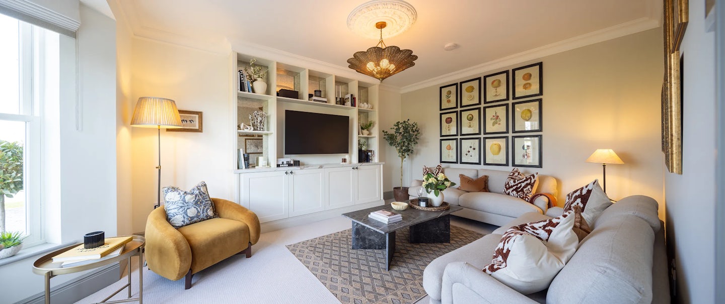 Living room with a modern design. A large built-in shelving unit frames a flat-screen TV, surrounded by books and decorative items. Two sofas—one beige, one light gray—are adorned with patterned cushions, complemented by a mustard-coloured armchair near a window. A coffee table sits atop a patterned rug, holding books and decor. Framed botanical prints in a grid pattern decorate the walls. Various lighting sources, including a ceiling fixture, floor lamp, and table lamp, create a warm ambiance. A potted plant adds greenery to the inviting space.
