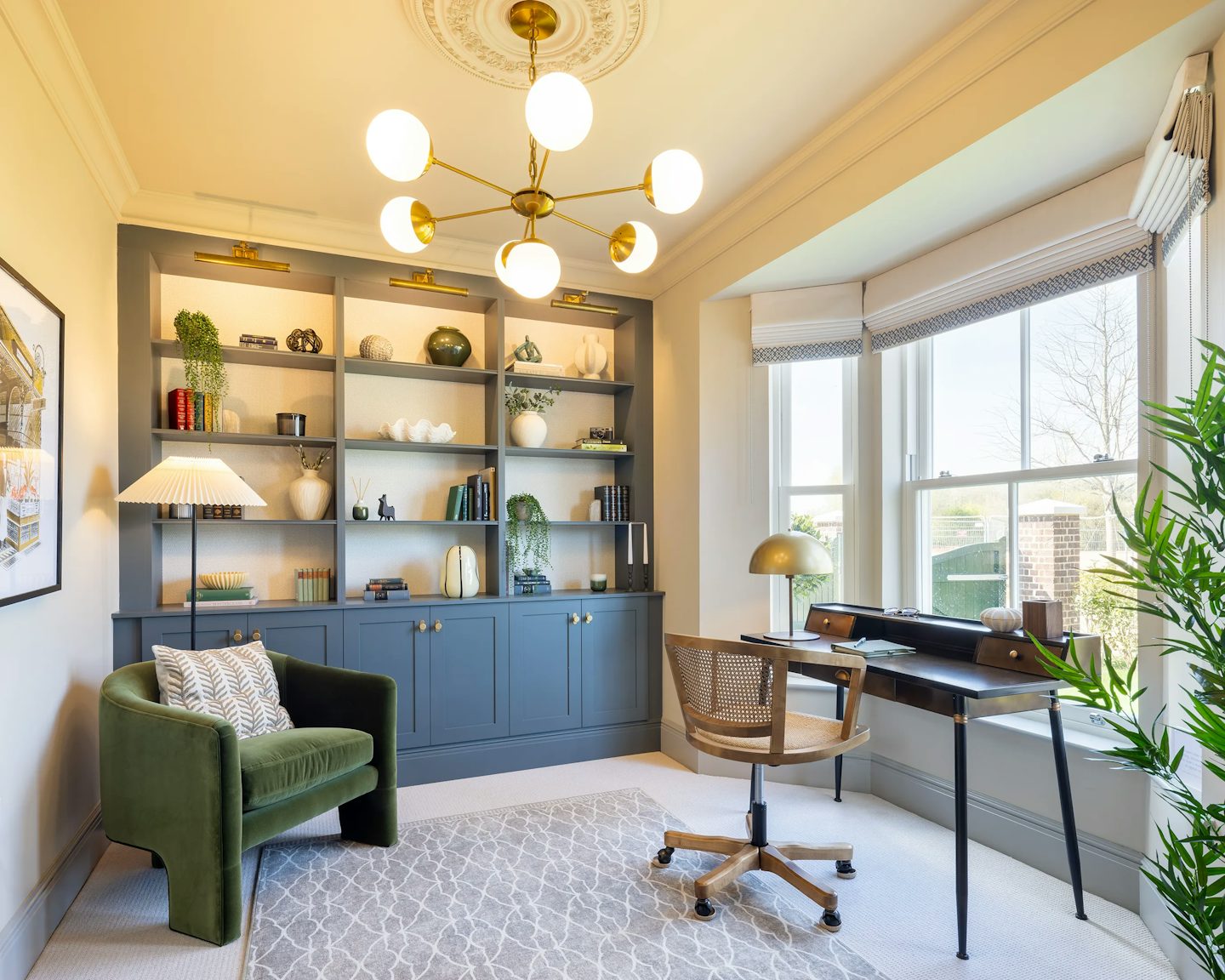 A well-decorated home office featuring a wooden desk with a black surface, a modern lamp, and a swivel chair. A green armchair with a patterned cushion sits nearby, next to built-in shelves filled with books, vases, and sculptures. The room is illuminated by a spherical chandelier and natural light from a large window with white blinds. A patterned rug adds warmth to the space.