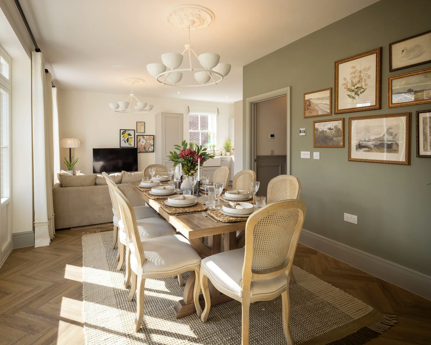 A bright and modern dining and living area with a warm, inviting atmosphere. The wooden dining table is set with plates, glasses, and a floral centrepiece, surrounded by six chairs atop a beige rug. In the adjacent living space, a beige sofa sits near a TV and a floor lamp, with framed artwork adorning the walls. Elegant chandeliers illuminate the room, complemented by natural light streaming in through the windows.