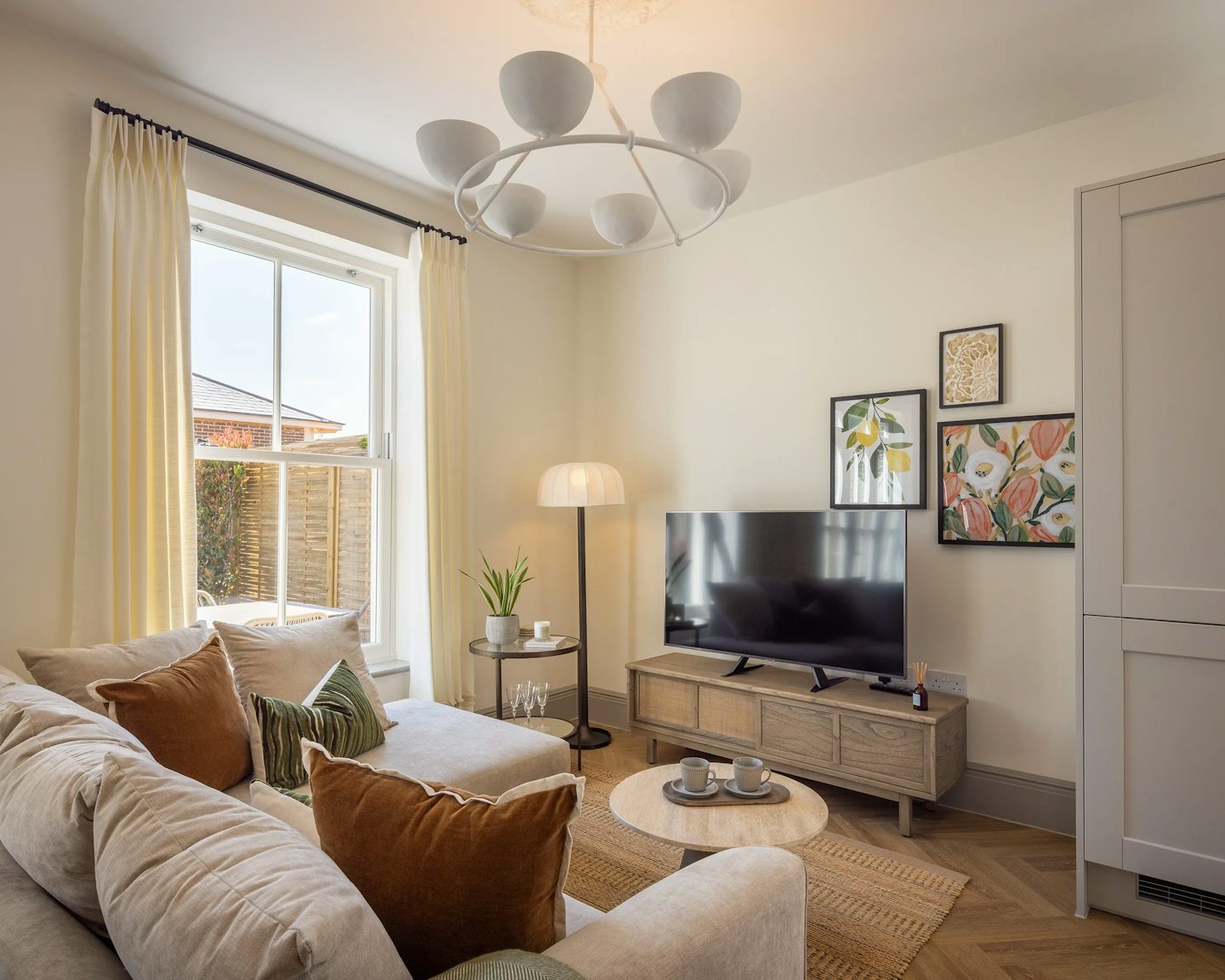 A cosy kitchen dining living area with a modern design. A large window with cream-colored curtains lets natural light in. A beige sectional sofa adorned with brown and green throw pillows sits around a round wooden coffee table with a decorative tray. A circular floor lamp stands nearby. Against the opposite wall, a wooden TV stand holds a flat-screen TV and decorative accents. Above the TV, three framed botanical artworks add a stylish touch. A unique chandelier with multiple circular lights hangs from the ceiling, enhancing the warm, inviting ambiance.