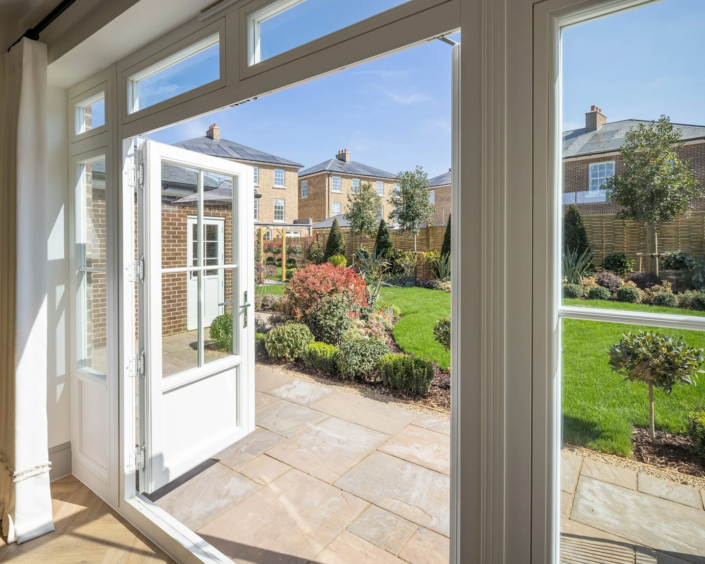 A view from inside a house looking out through glass doors onto a well-maintained garden. The garden features a variety of plants, shrubs, and small trees, with neatly trimmed hedges and a paved patio area. In the background, several brick houses with chimneys and large windows suggest a residential neighborhood. The scene is bathed in sunlight, creating a warm and inviting atmosphere.