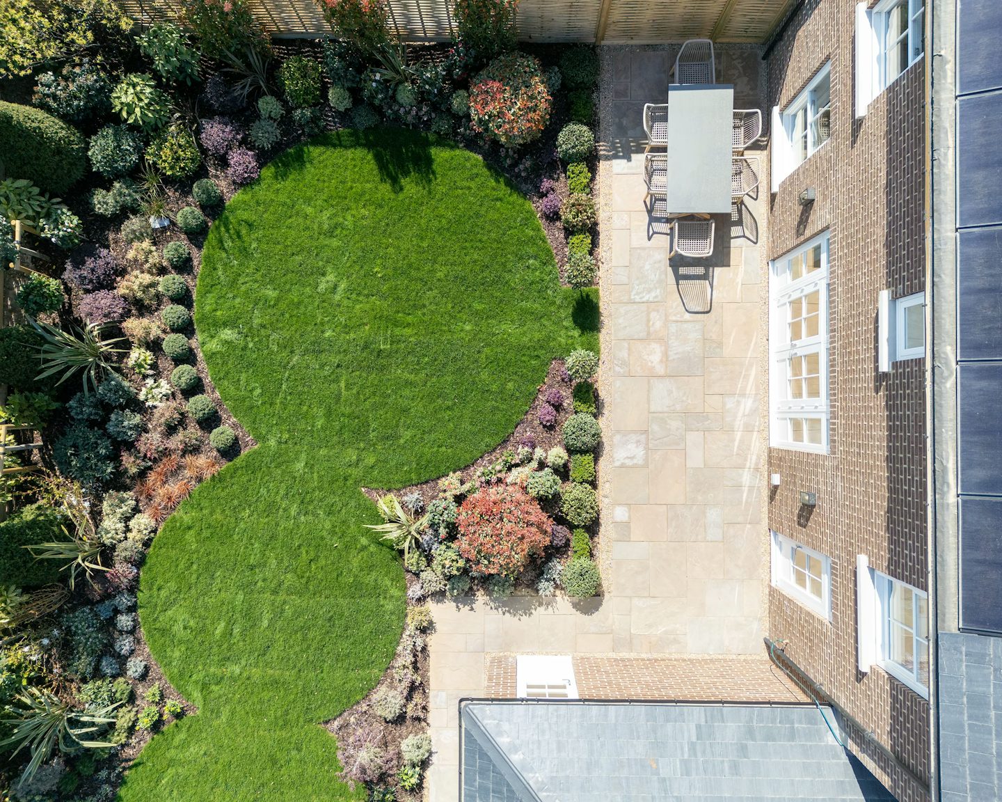 An aerial view of a beautifully landscaped backyard garden. The space features a neatly trimmed lawn with two large, circular grassy areas. Surrounding the lawn, various plants and shrubs are arranged in a landscaped pattern, adding depth and texture. A paved patio area near the house includes a rectangular table with four chairs, creating an inviting outdoor seating area. The house, with multiple windows overlooking the garden, is framed by a wooden fence that encloses the space. The combination of greenery, vibrant flowers, and structured design highlights a well-maintained and visually appealing garden.