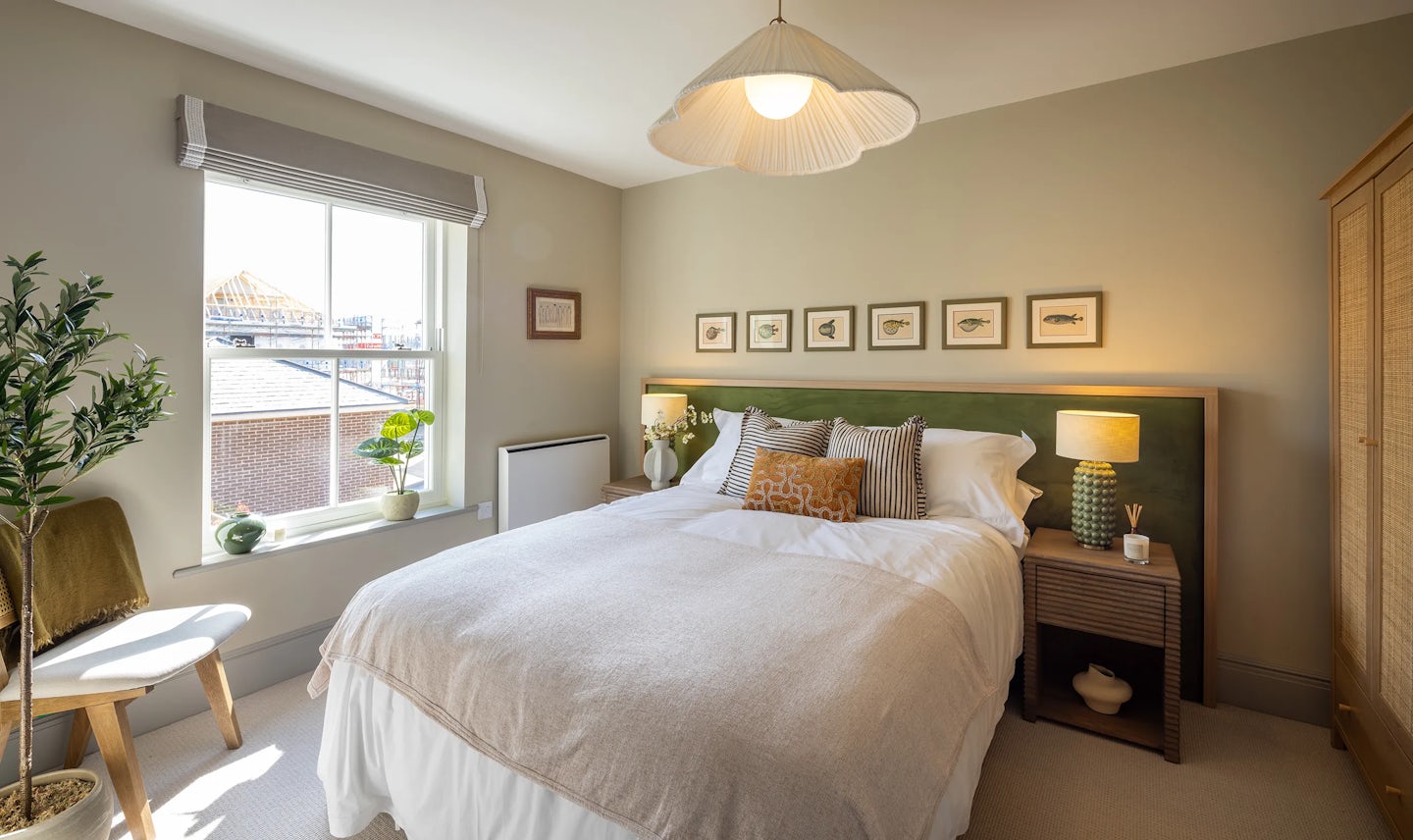 Bedroom with a double bed dressed in white bedding and multiple pillows, including a patterned throw pillow. Above the bed, six framed fish illustrations add a decorative touch. To the right, a wooden nightstand holds a lamp, a small vase, and a book. On the left, a window lets in natural light, showing neighboring buildings, with a potted plant on the windowsill and a chair draped with a blanket nearby.
