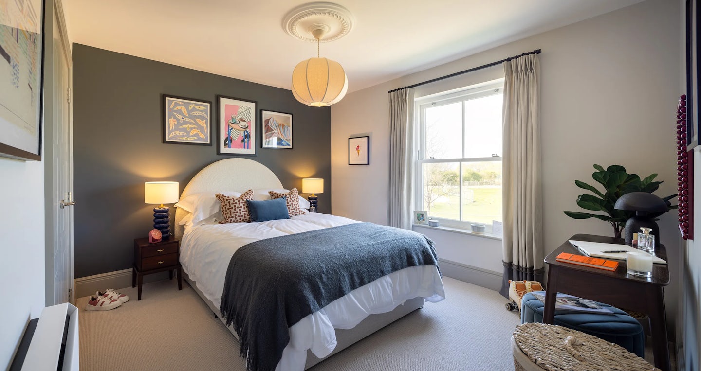 Bedroom featuring a bed with white linens and a dark grey blanket, positioned against a dark accent wall with three framed artworks. Two bedside tables with lamps flank the bed. To the right, a large window with beige curtains lets in natural light. A small desk with a lamp, books, and a potted plant sits near the window.
