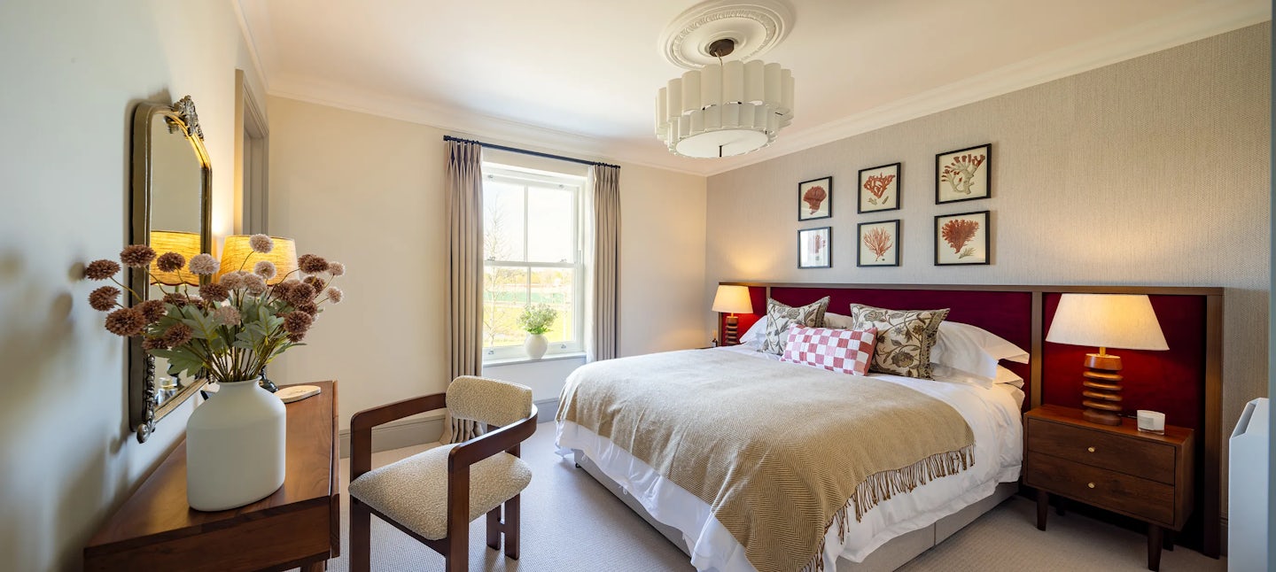 A cozy bedroom featuring a neatly made double bed with a beige blanket, white sheets, and an assortment of patterned pillows, including floral and checkered designs. Above the bed, six framed botanical prints are arranged in two rows. Wooden nightstands on either side hold lamps, complementing the room’s warm lighting. A large window with beige curtains allows natural light to fill the space. A wooden dresser with a mirror and a vase of dried flowers adds to the decor, while a cushioned chair is placed nearby for seating.