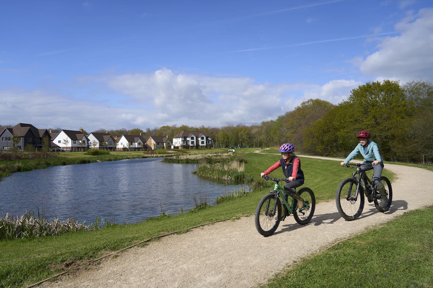 Two children ride bicycles along a gravel path beside a calm lake. The path is lined with lush green grass and trees, and Thakeham built houses are visible in the background. The sky is blue with scattered clouds.