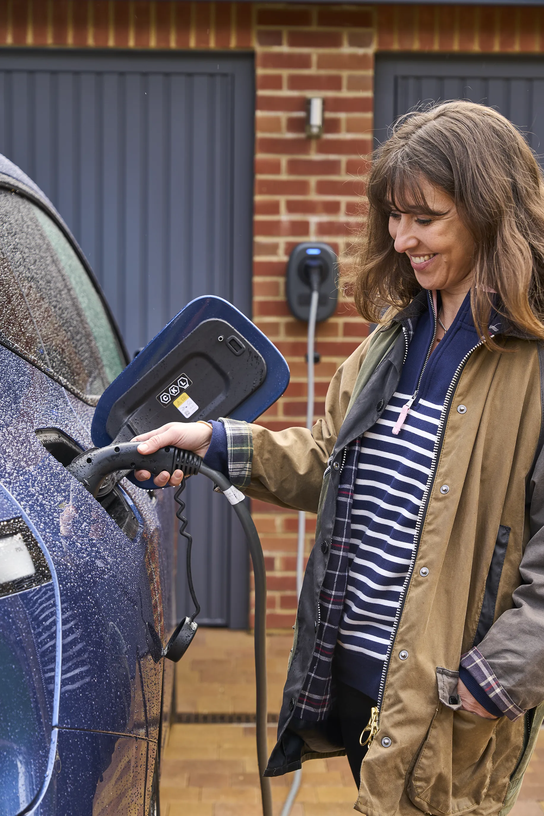 A woman plugs a charging cable into the port of an electric vehicle. The charging station is mounted on a brick wall with two closed garage doors in the background.