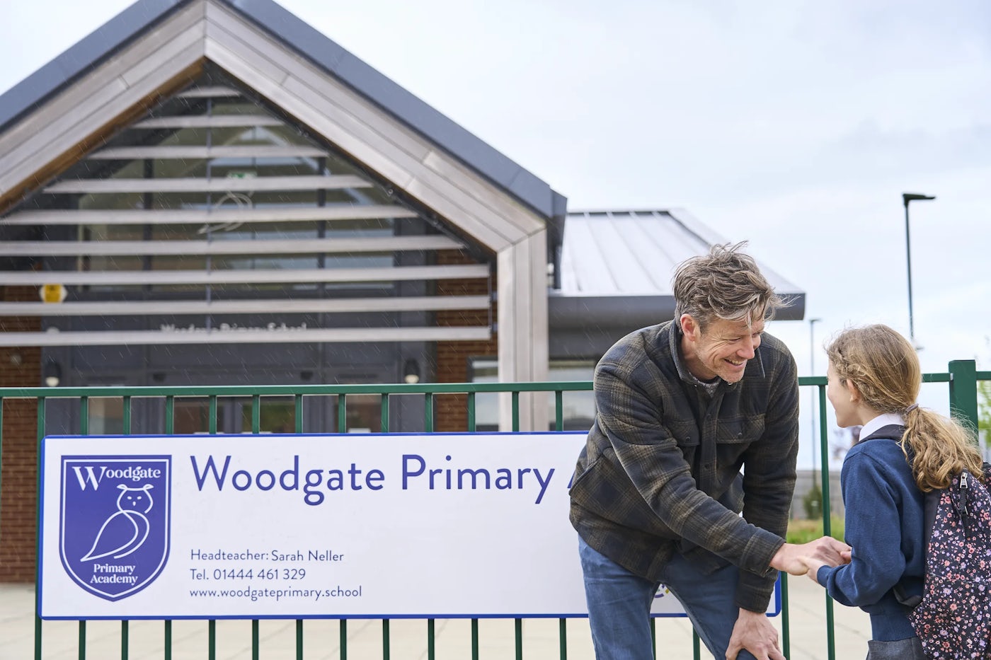 The entrance of Woodgate Primary Academy features a sign displaying the school's name, logo, headteacher's name, contact information, and website. In the foreground, a father bends down to speak with his daughter, who is wearing a school uniform and carrying a backpack. The background showcases the school’s modern building with a pitched roof and large windows.