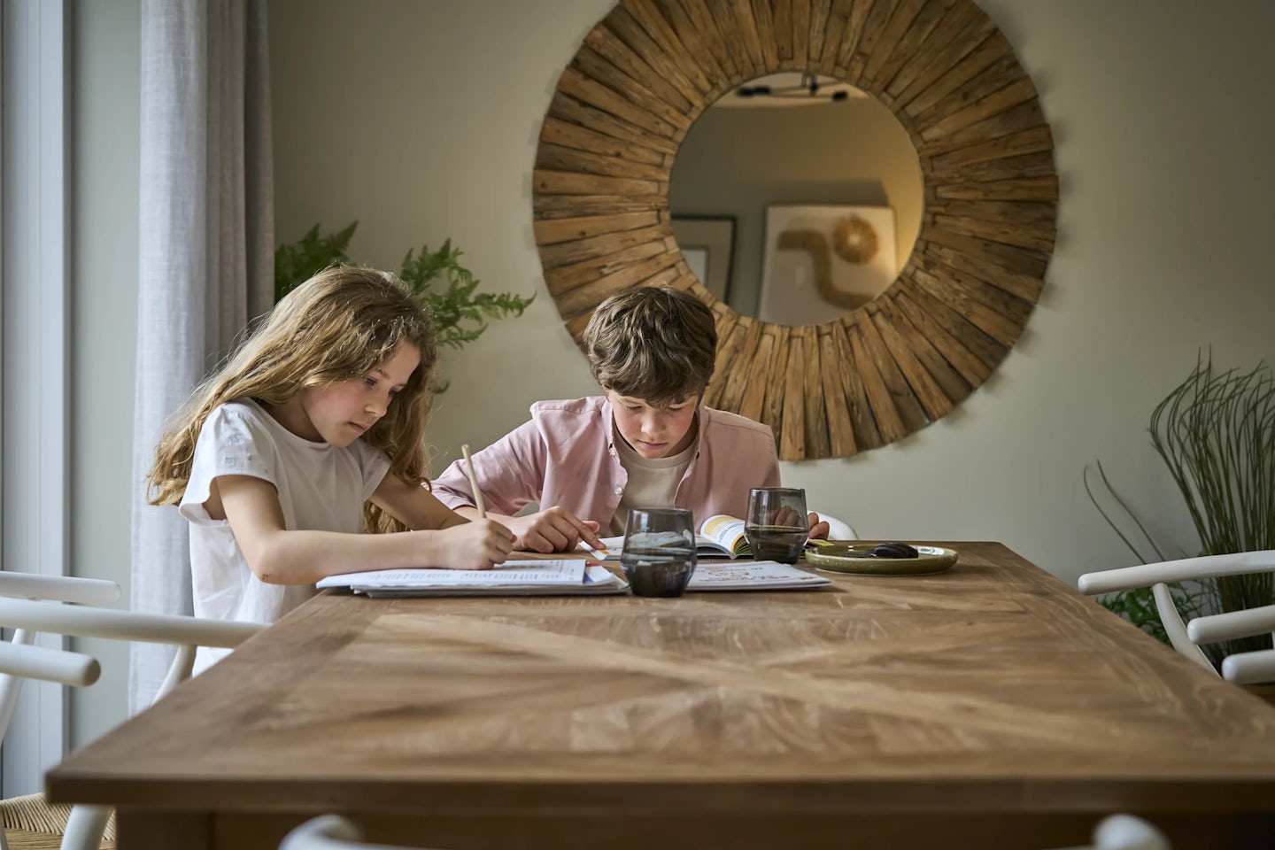 Two children sit at a wooden table, focused on their studies. The table has a natural wood finish, with two glasses and a plate holding some items. Behind them, a large circular mirror with a wooden frame reflects part of the room. The neutral-toned space features light walls and a plant in the background, creating a calm atmosphere.